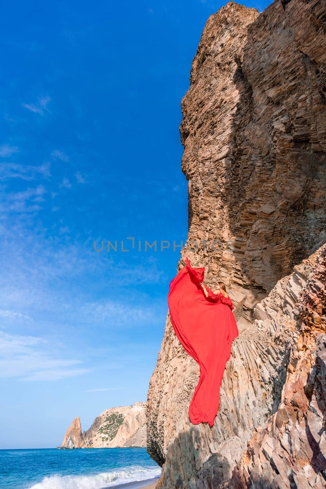 woman sea red dress. Woman with long hair on a sunny seashore in a red flowing dress, back view, silk fabric waving in the wind. Against the backdrop of the blue sky and mountains on the seashore