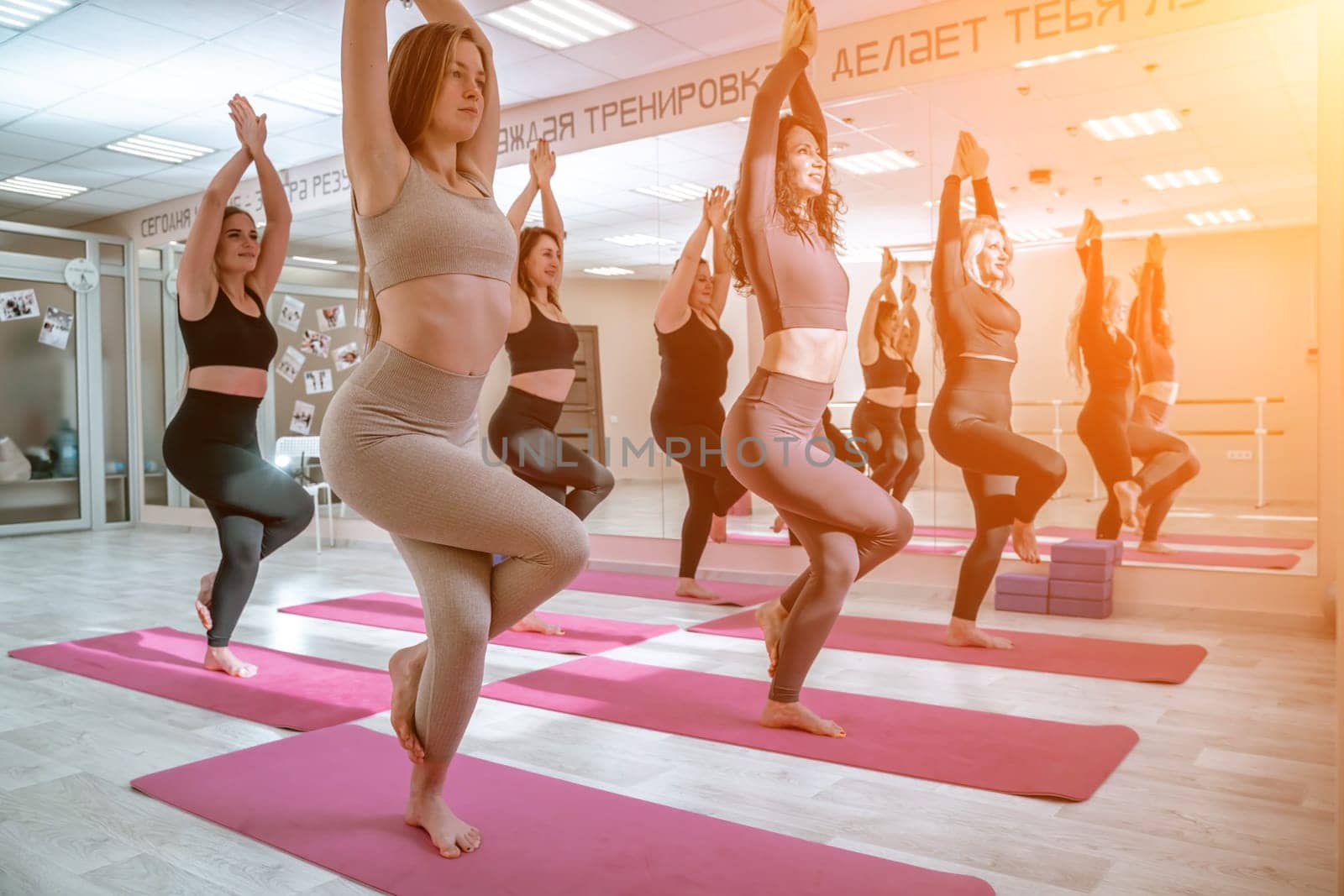 A group of six athletic women doing pilates or yoga on pink mats in front of a window in a beige loft studio interior. Teamwork, good mood and healthy lifestyle concept