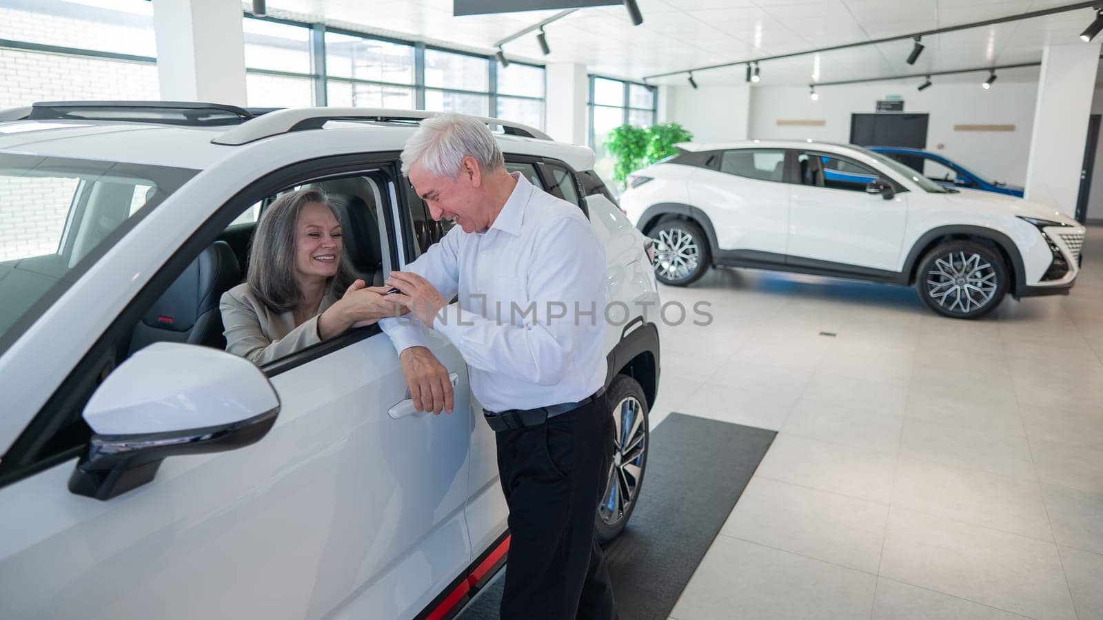 Mature Caucasian woman sits in a new car, an elderly man gives her the keys. by mrwed54