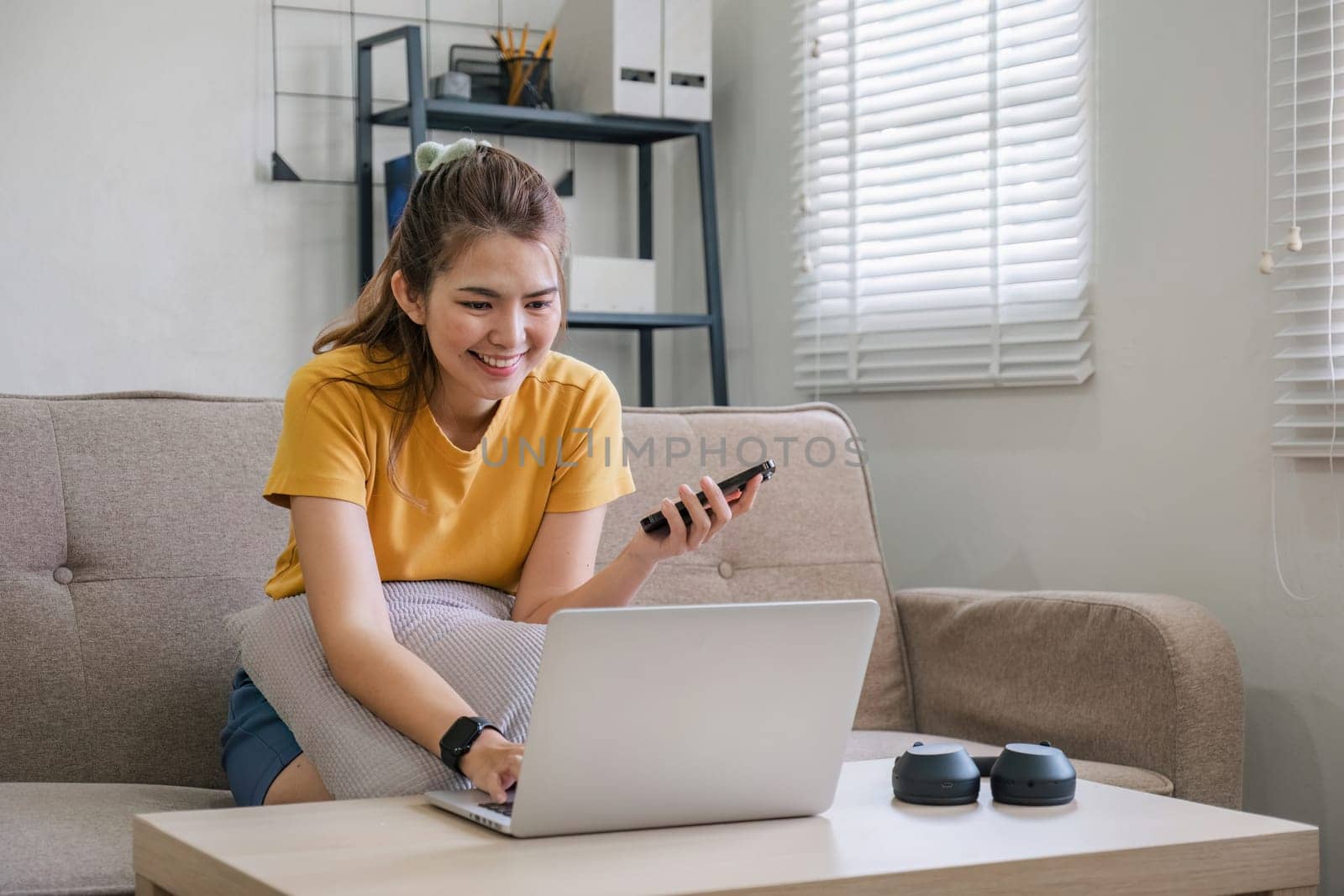 Happy young Asian woman uses mobile phone while sitting on sofa at home with laptop computer..