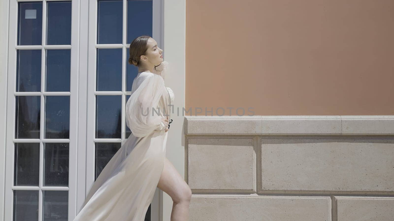 Side view of a gorgeous female model in a silk dress swaying in the wind. Action. Woman standing and posing in front of a building with big window and brown facade