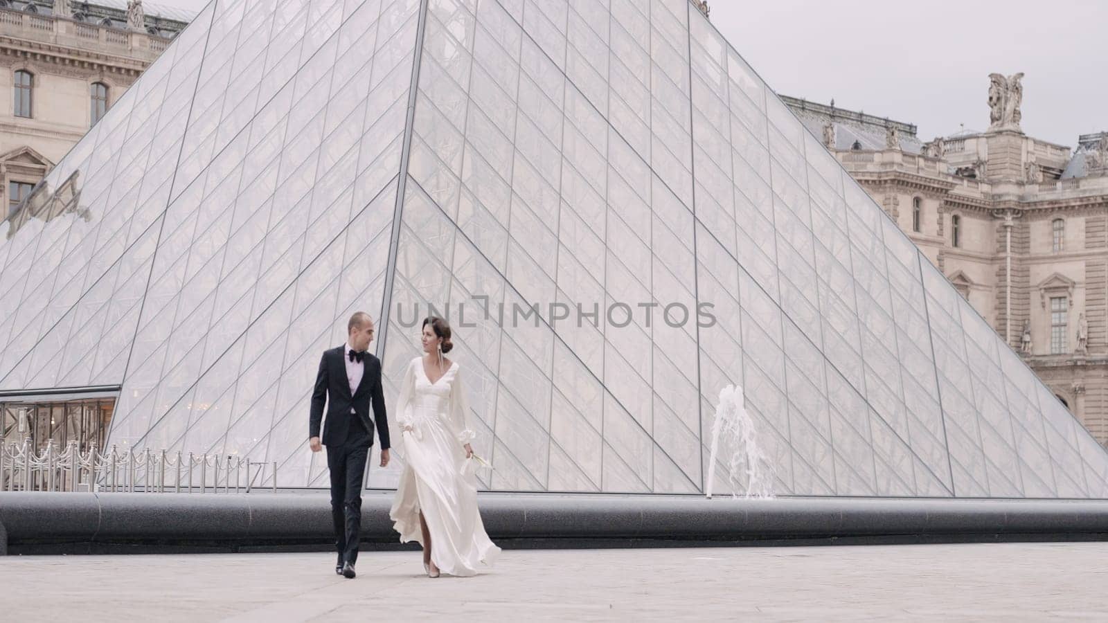 Elegant couple walks in ancient square. Action. Beautiful happy couple in luxurious outfits are walking in ancient city. Couple in love in elegant outfits walk around Louvre.