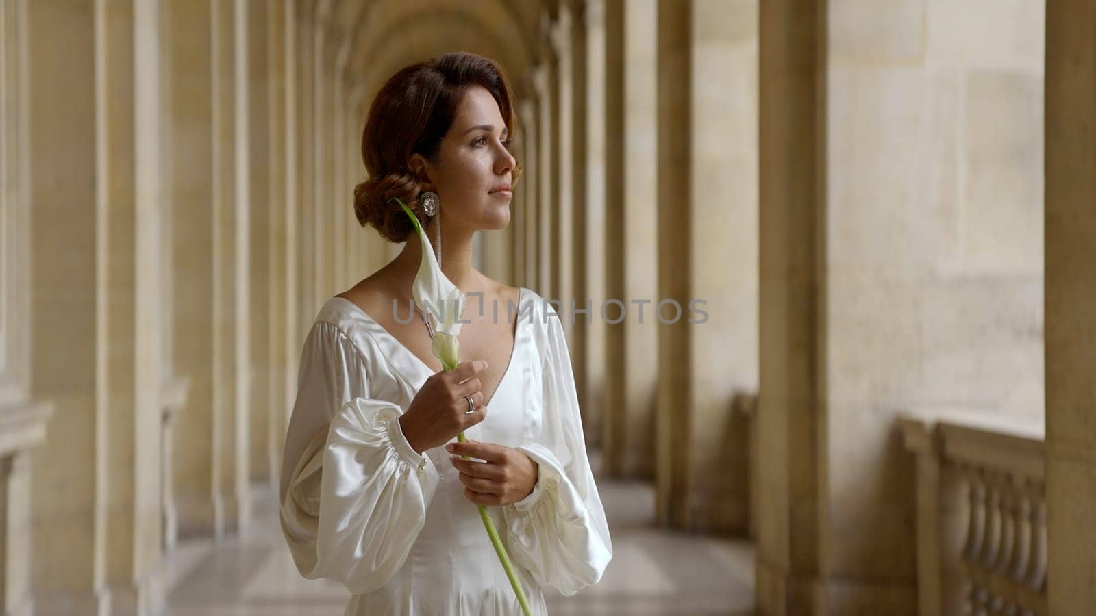 Beautiful young romantic woman in a long white dress standing near ancient columns. Action. Beautiful woman holding a flower in her hand . by Mediawhalestock