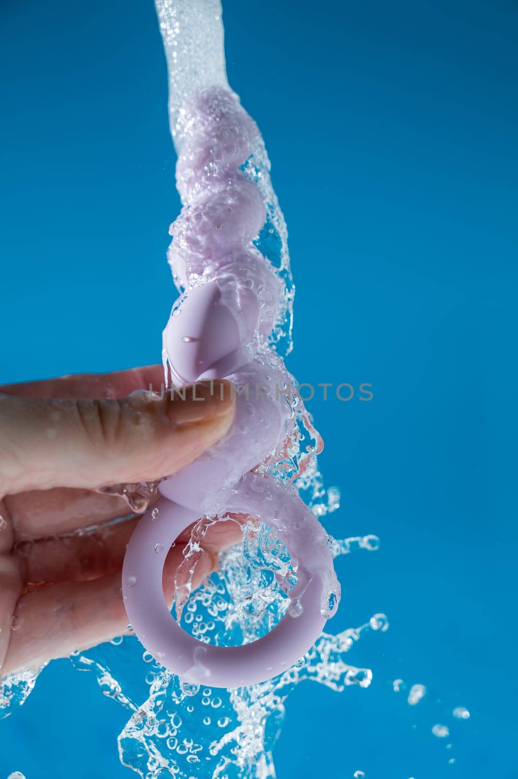 Woman holding lilac anal beads under running water on blue background. Sex toy hygiene concept. by mrwed54