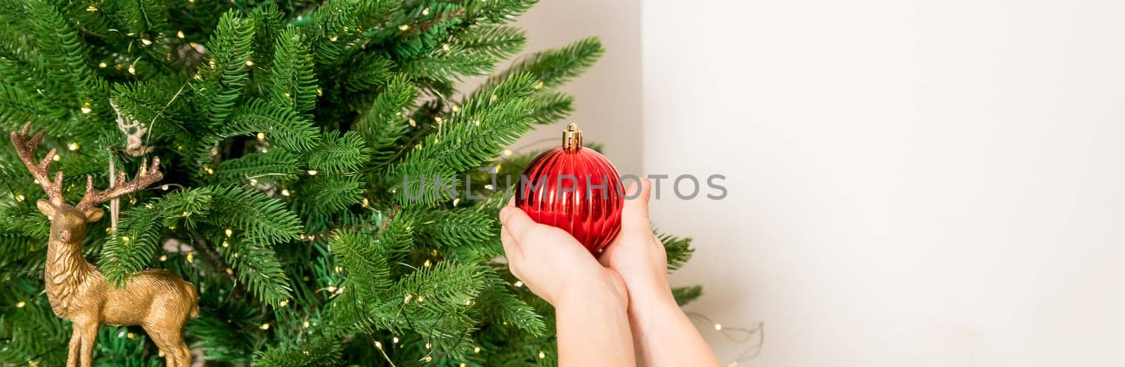 winter holidays and people concept - close up of child hand decorating tree with red ball.Anonymous little girl decorates christmas tree.Cozy magical atmosphere in the room. copy space. web banner by YuliaYaspe1979