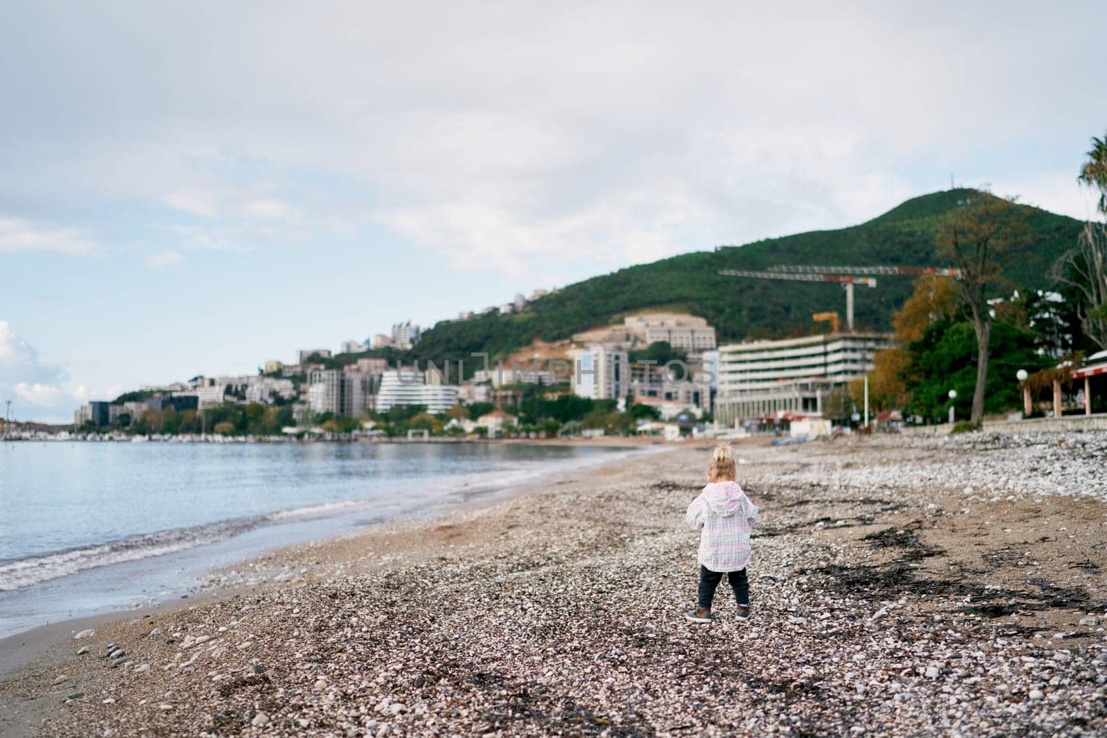 Little girl walks along the pebbly seashore against the backdrop of mountains and apartment buildings. Back view. High quality photo