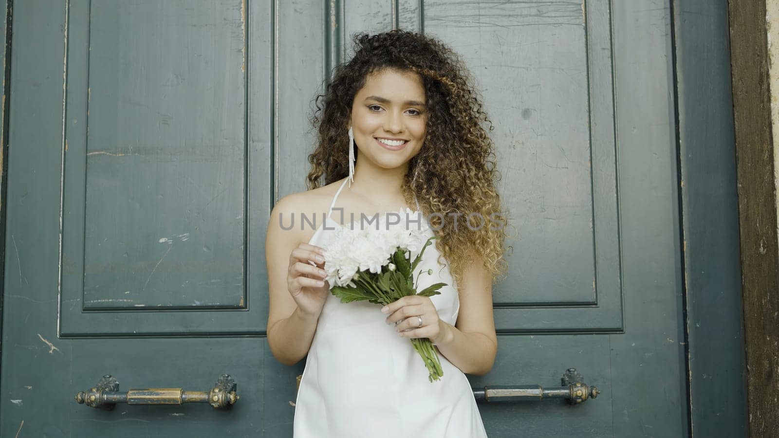 Gorgeous woman in white dress with curly hair with bouquet in her hands against the dark green door. Action. Bride waiting for the groom