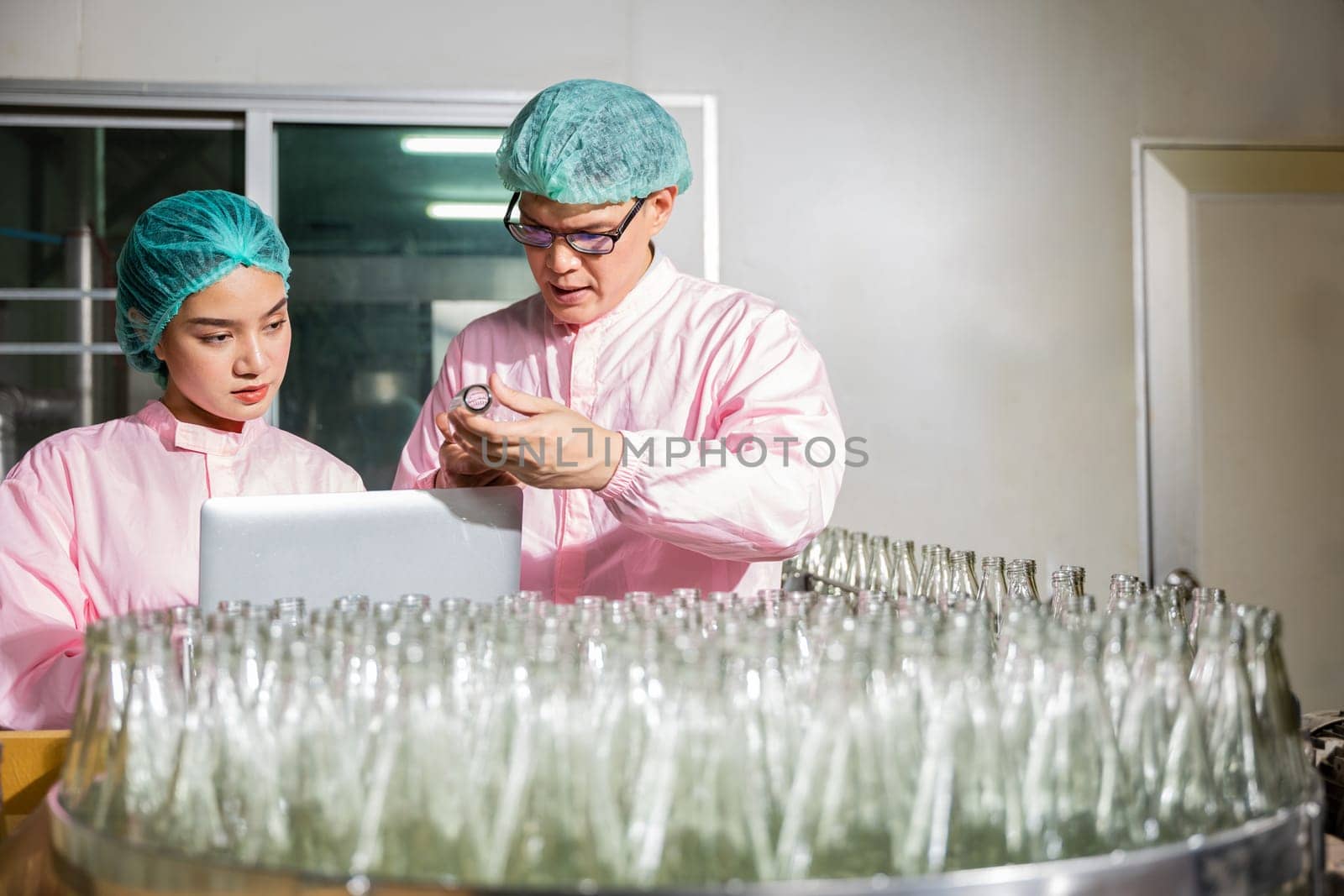 In a beverage factory line two engineers meticulously check product bottles on the conveyor belt. Quality control and inspection managed by an engineer and inspector using a laptop.