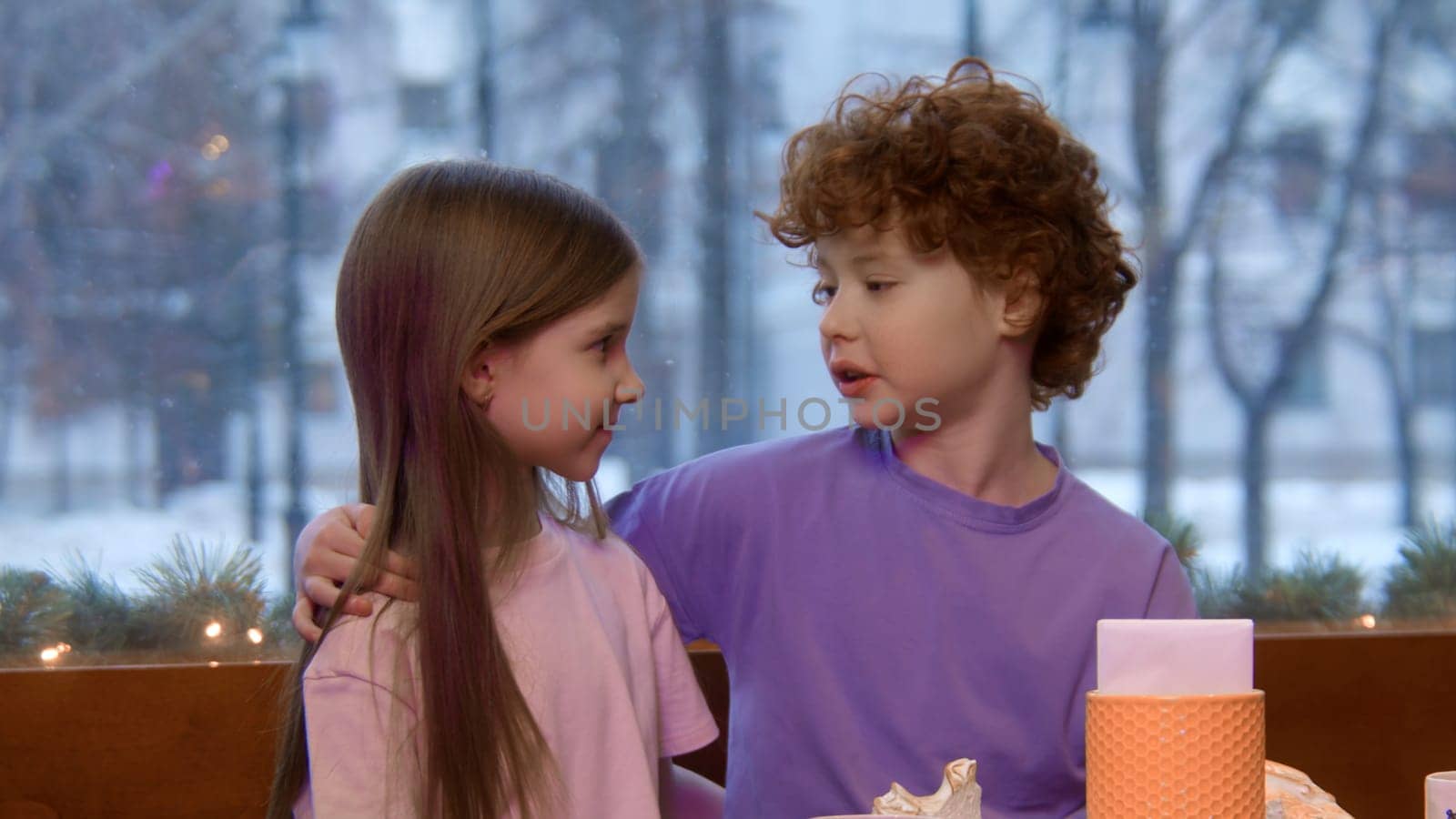 Children sitting together at a table in a restaurant. Stock footage. Brother with red curly hair putting ihs arm around the shoulders of her cute sister