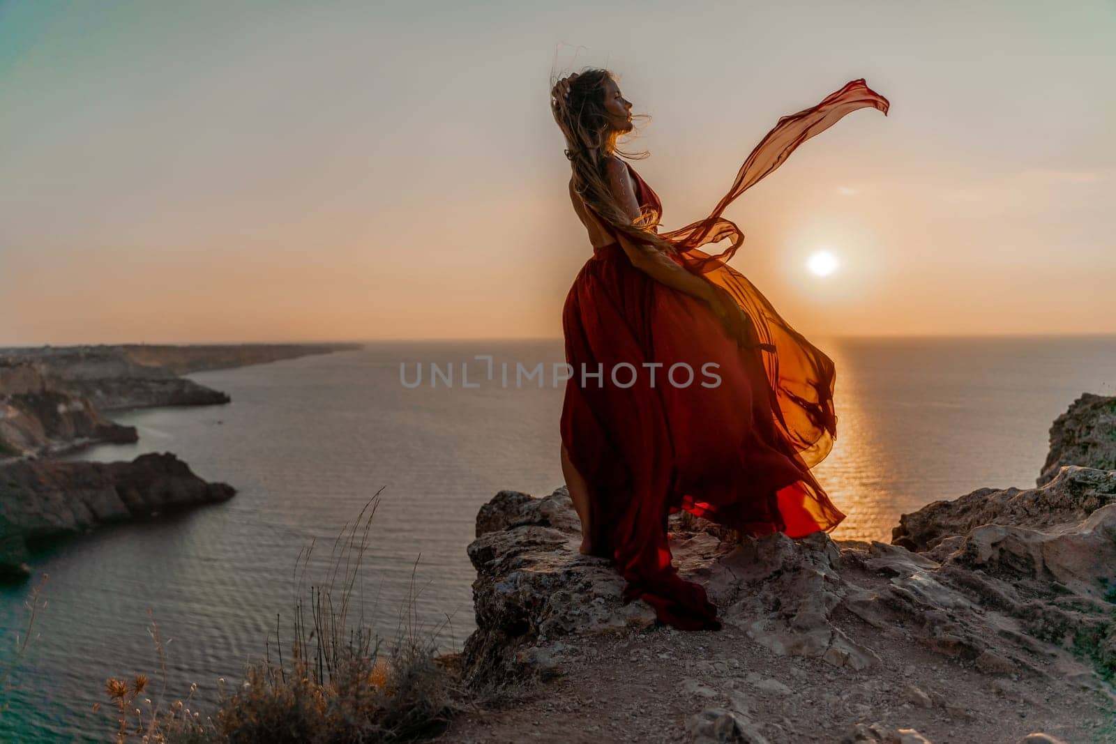 Woman sunset sea red dress, side view a happy beautiful sensual woman in a red long dress posing on a rock high above the sea on sunset