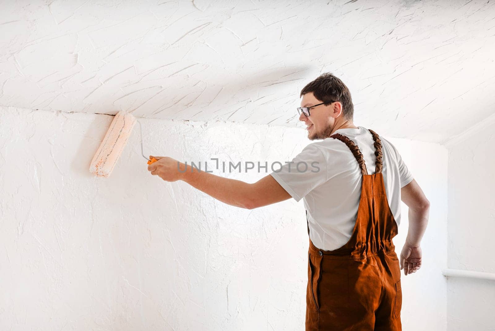 Color of the year 2024: Peach Fuzz. Repair, home improvement and renovation concept. young man in overalls painting wall into color with a rolle