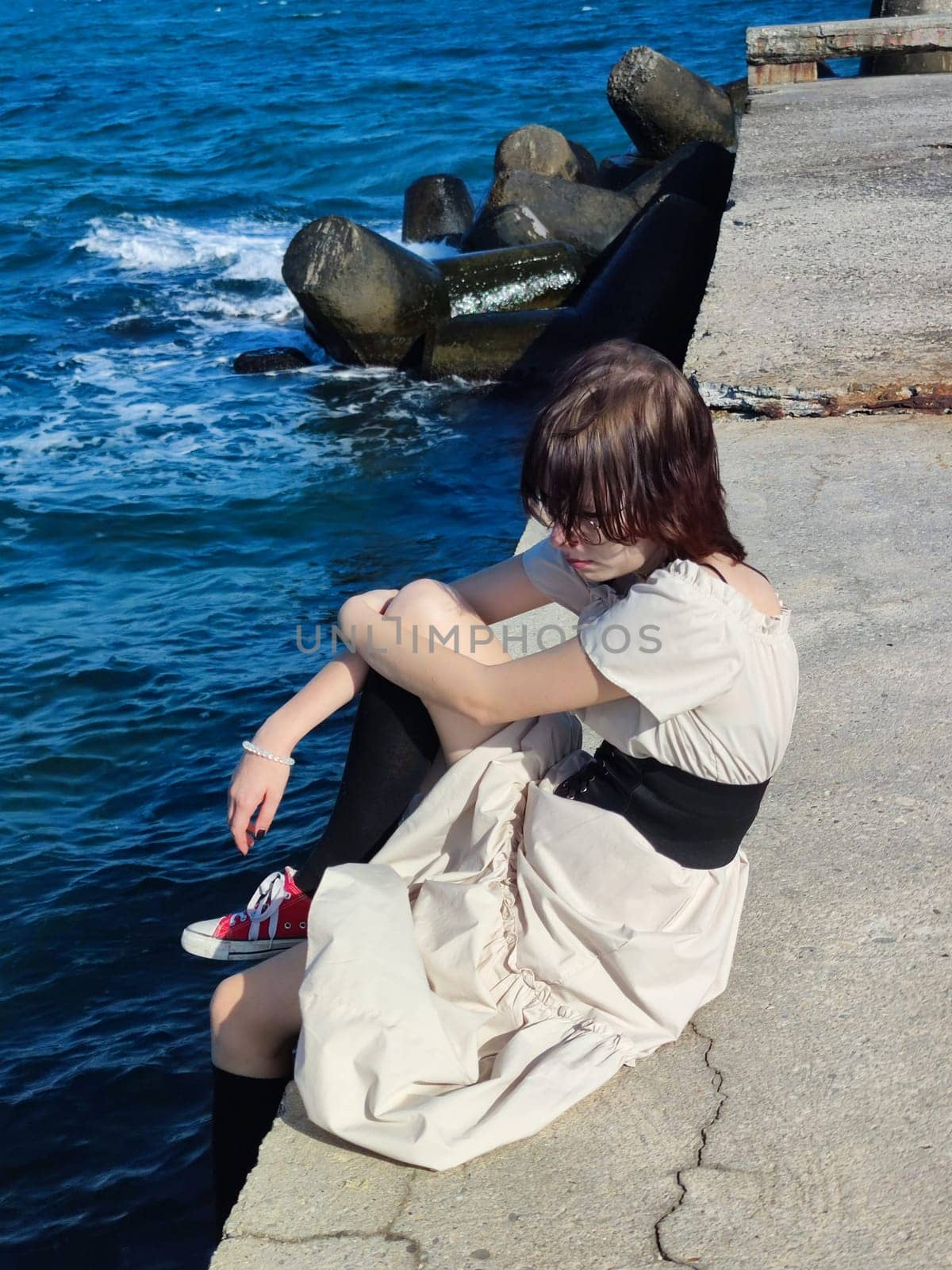 sad teenage girl with short dark hair in a white dress sits thoughtfully on the sea pier.
