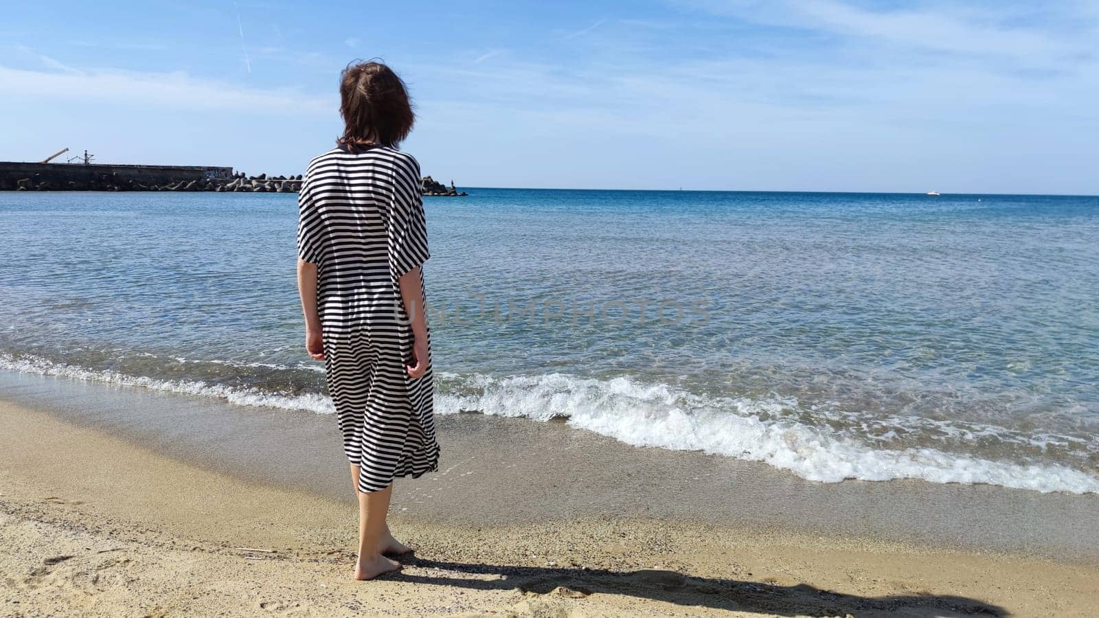 Barefoot teenage girl in a striped dress on the seashore on a sunny day, rear view