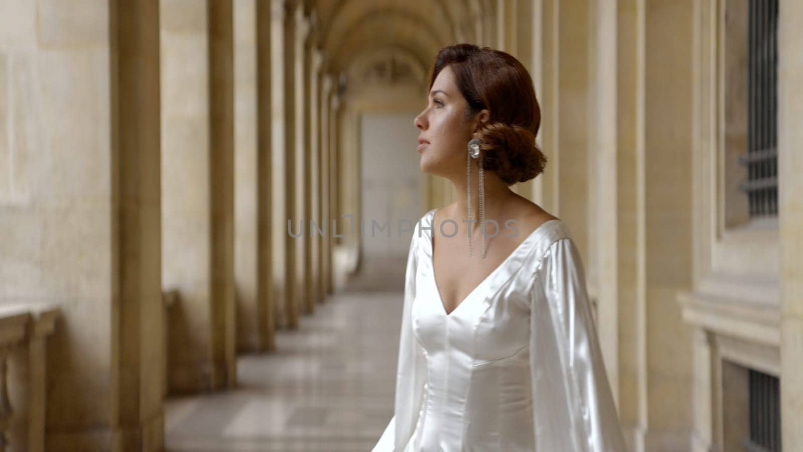 Woman in romantic white silk dress looking in the distance on the background of an ancient building. Action. Elegant woman with a long earring. by Mediawhalestock