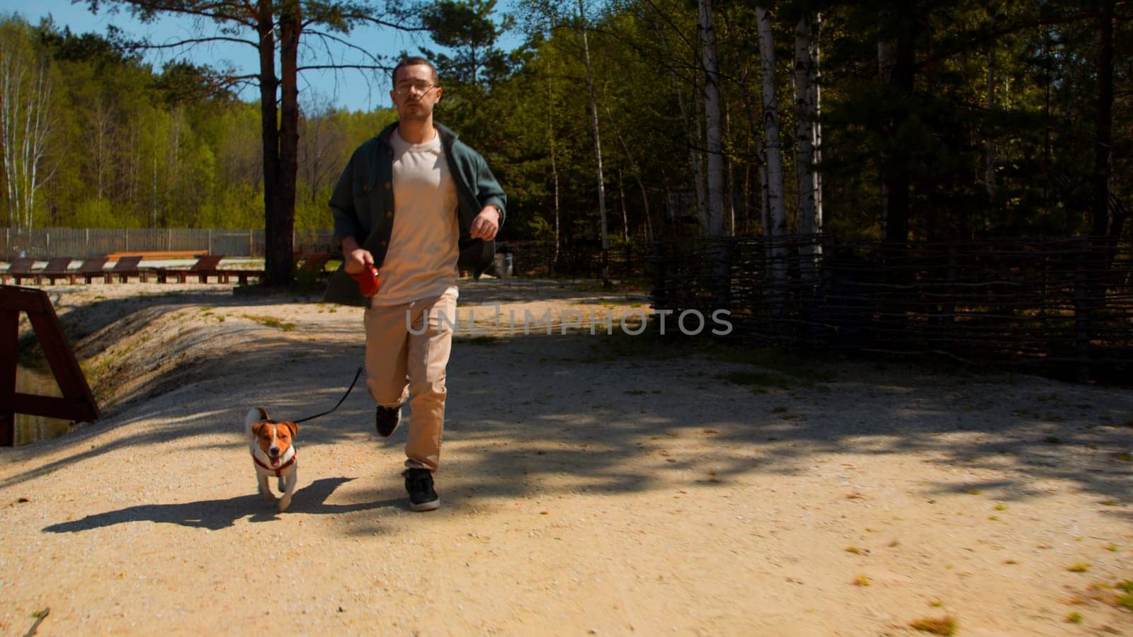 Happy man and his little cute puppy running along the green small lake. Stock footage. Man jogging with jack russell