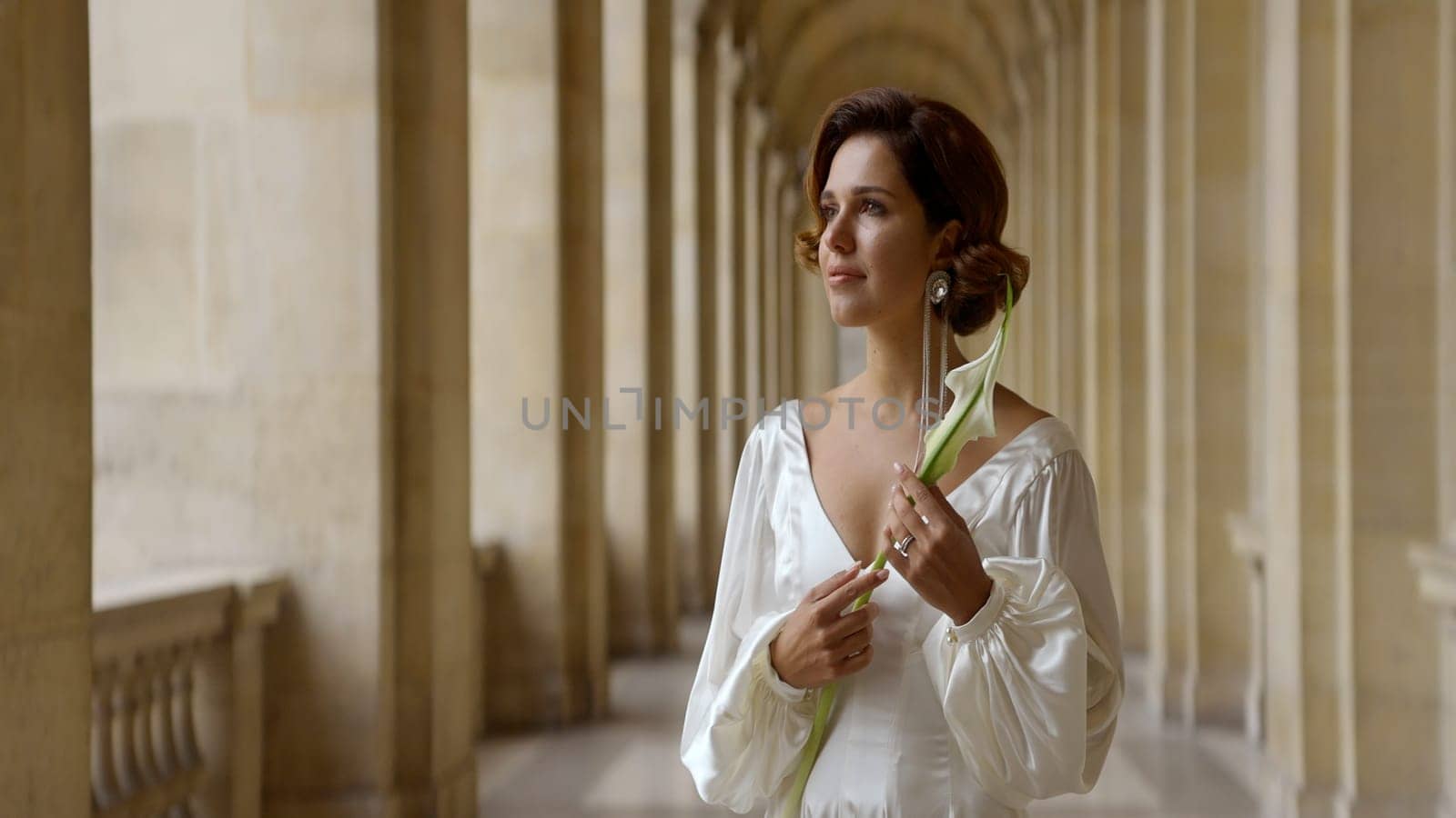 Beautiful young romantic woman in a long white dress standing near ancient columns. Action. Beautiful woman holding a flower in her hand . by Mediawhalestock