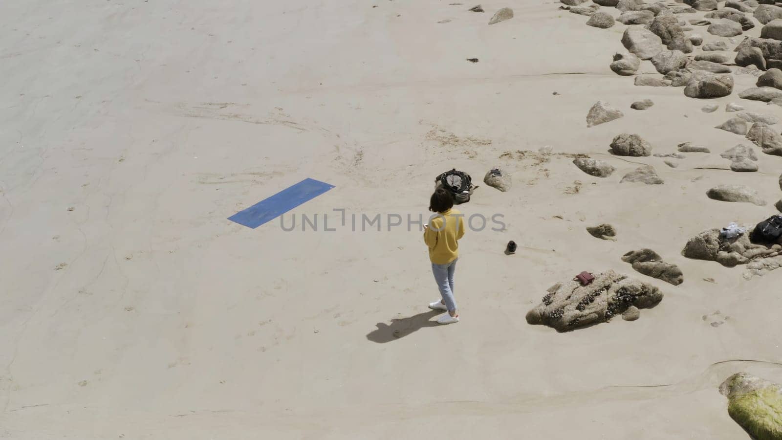 Aerial view of a woman wearing yellow sweater and blue jeans standing on the sandy sea shore near her yoga mat. Female standing on the sand near the stones.