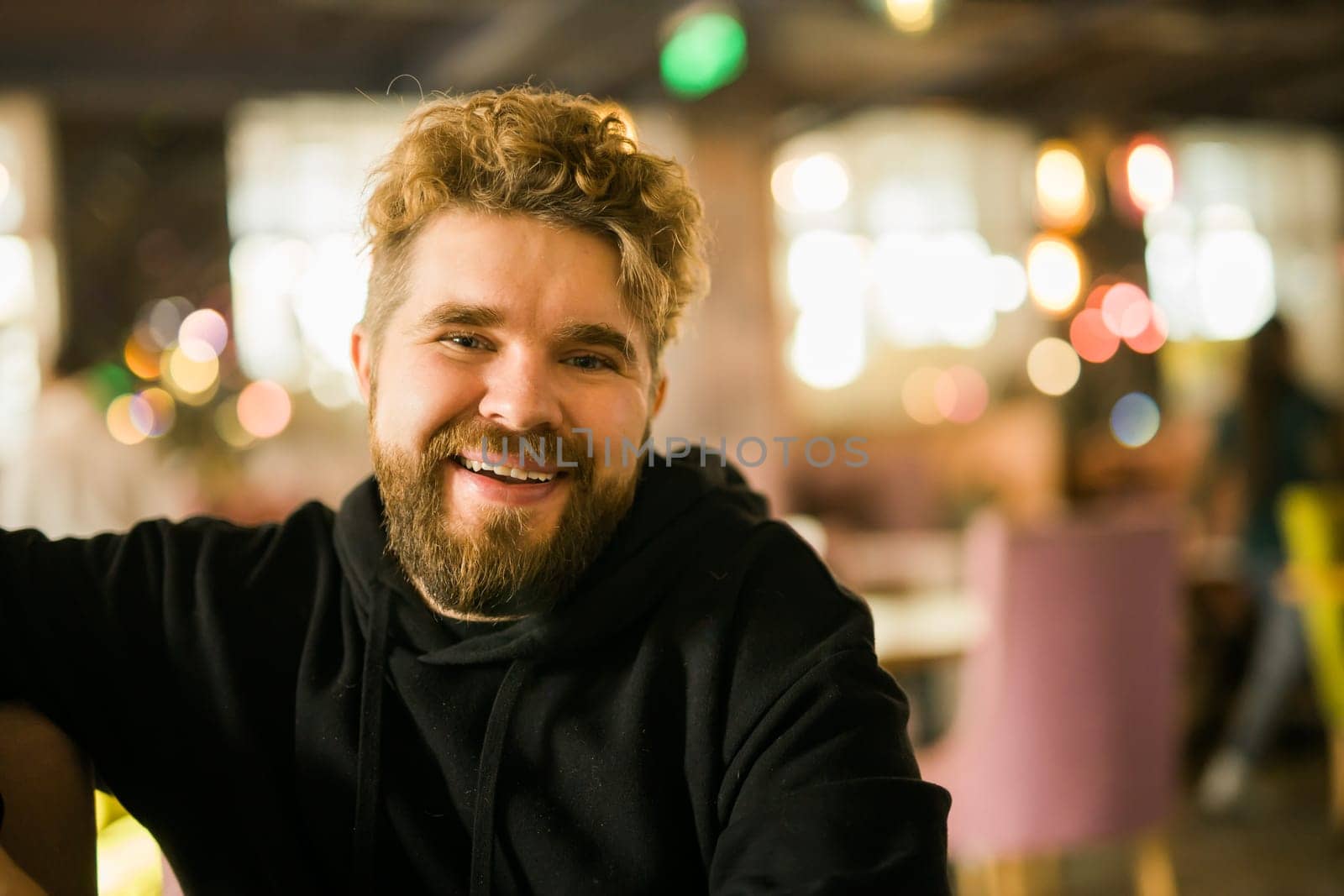 Bearded guy programmer or an entrepreneur in glasses in a stylish cafe looks at the camera and smiles copy space and empty space for advertising. Freelancer works remotely. Online communication video call and small business. by Satura86