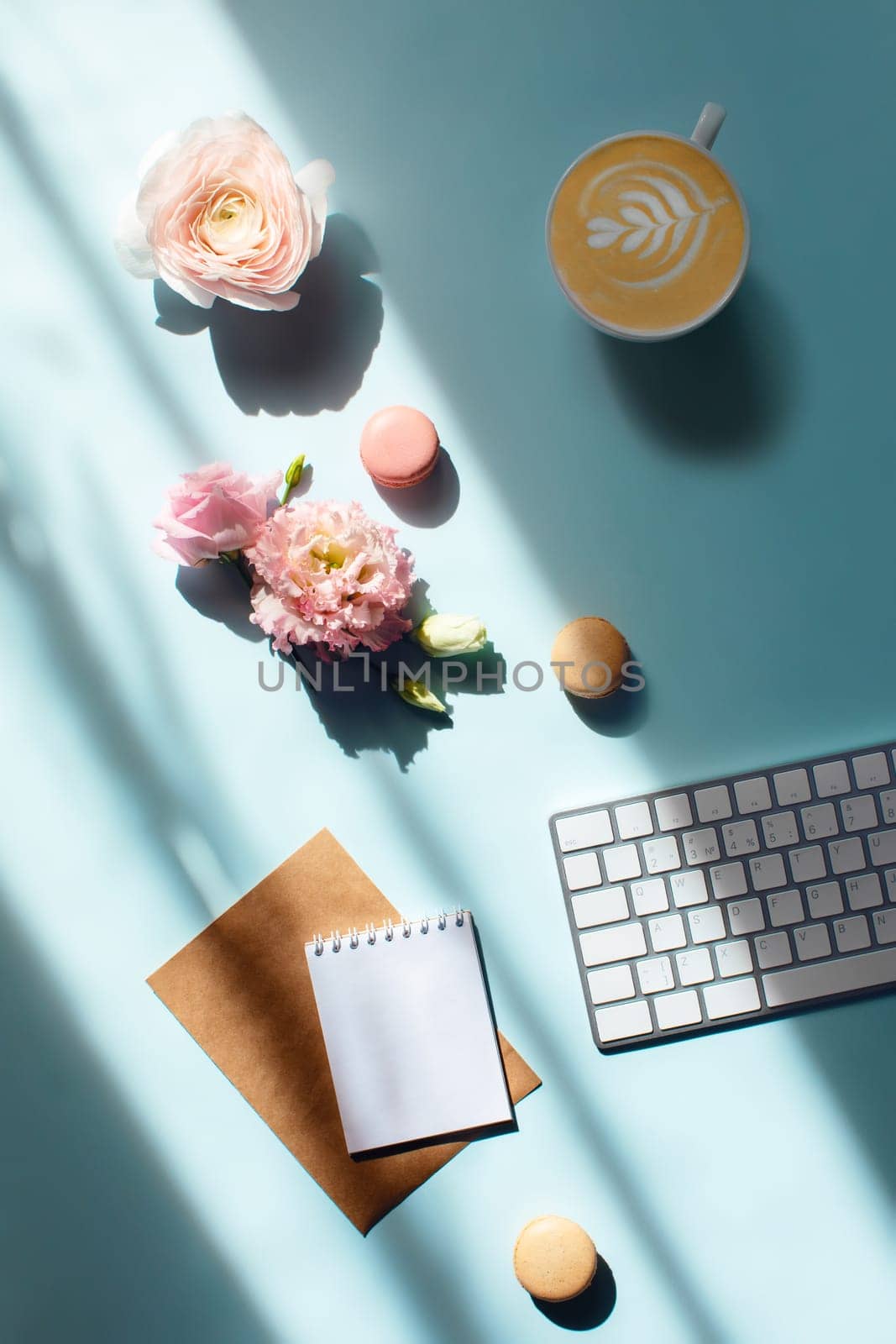 Vertical flat lay with tasty cappuccino and macaroons in sunlights with flowers and an envelope with a letter. Mental health support with aromatherapy and sweet food care while working at the computer.