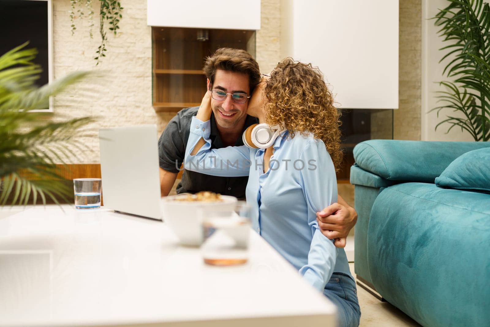 Cheerful young curly haired wife in smart casual clothes, hugging and kissing cheek of husband while bonding together and working on laptop in cozy living room