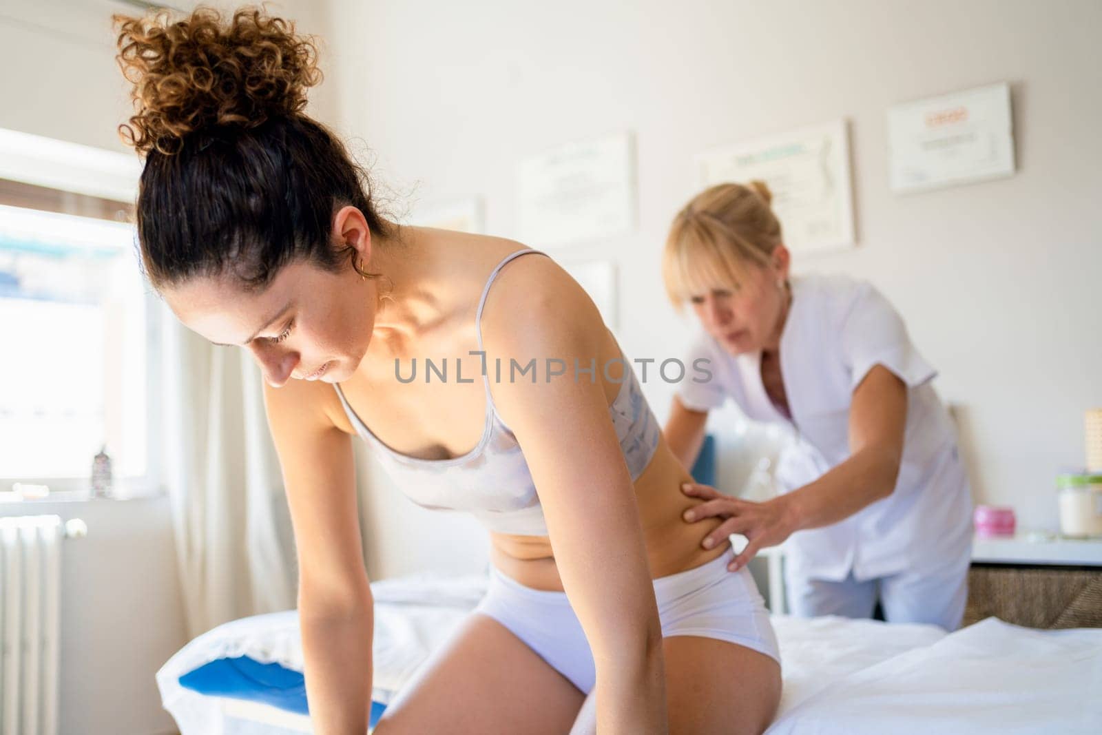 Concentrated masseuse looking down, while standing near spa bed and massaging lower back of young slim female sitting and bending forward during physiotherapy in osteopathy clinic