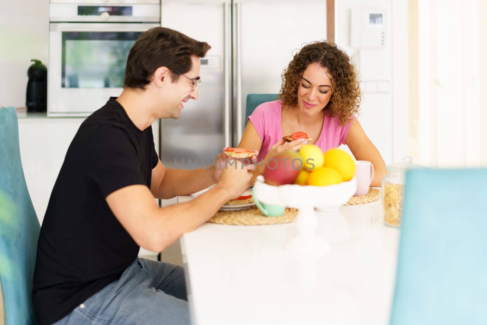 Smiling young couple sitting at table and enjoying healthy food in kitchen by javiindy