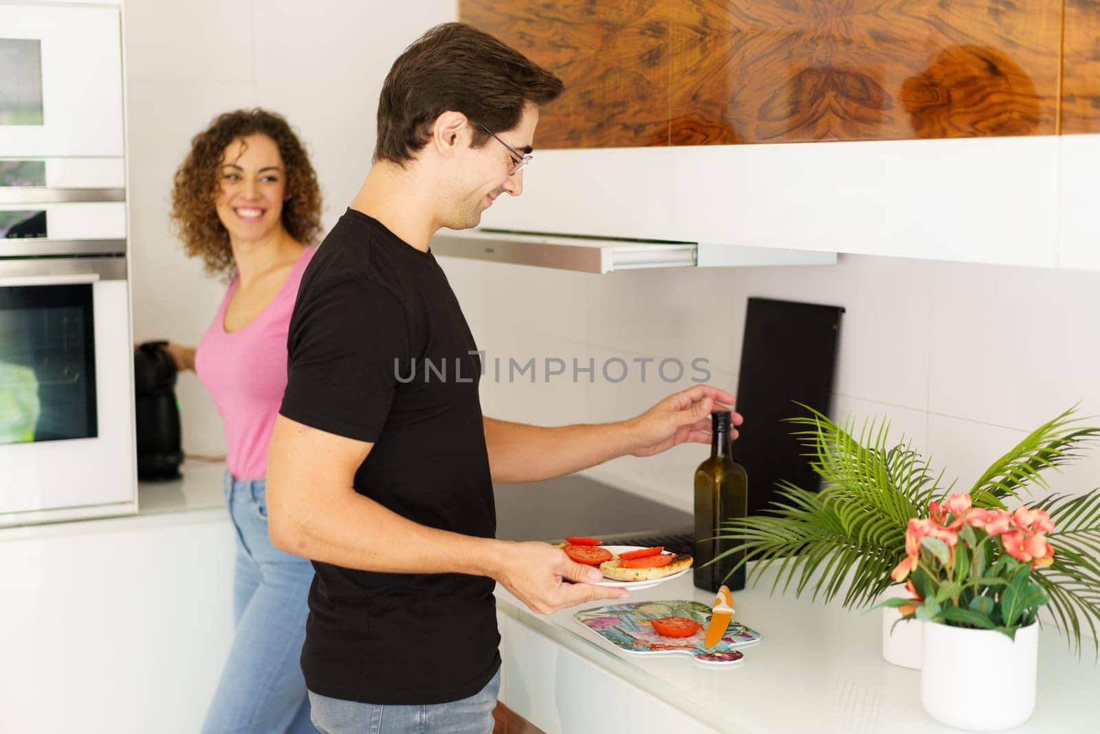 Side view of happy adult male looking down while standing with smiling conversing female at counter and carrying healthy salad, while opening wine bottle near flower pot in kitchen in daylight