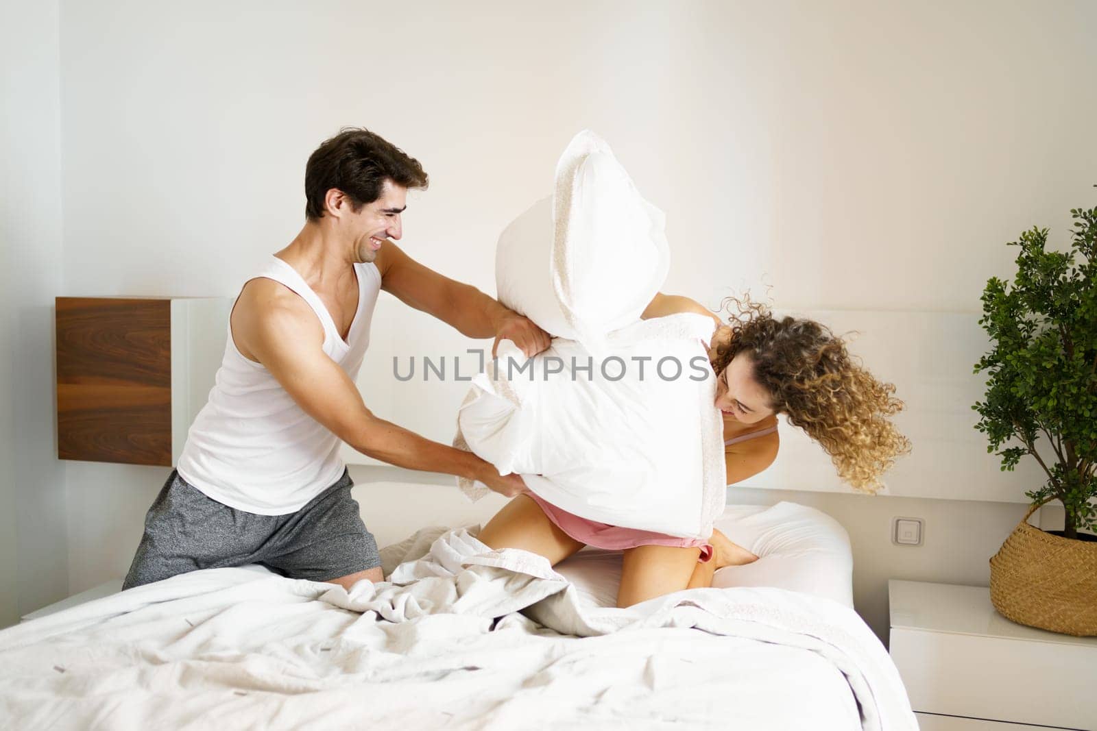 Side view of smiling adult male in nightwear looking away while kneeling on bed with eyes closed curly haired female, in bedroom and having fun with pillows fighting in daylight