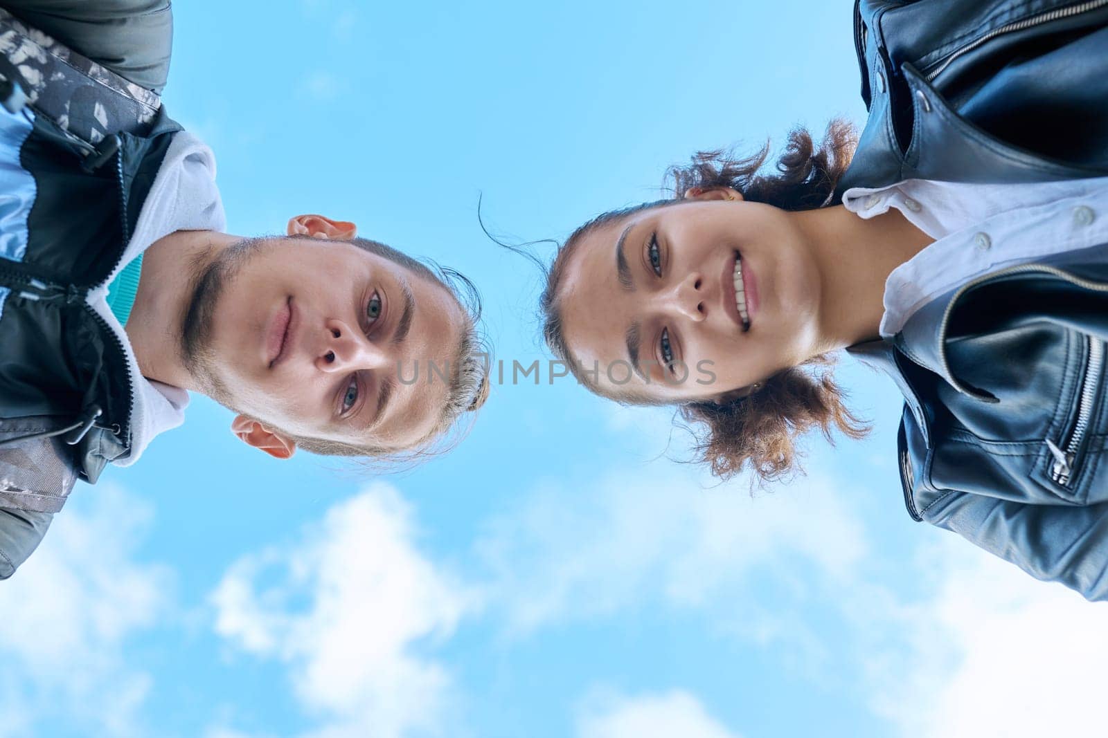 Closeup faces of young teenage guy and girl smiling looking down at camera, blue sky with clouds background. Youth communication leisure friendship lifestyle, college high school students education