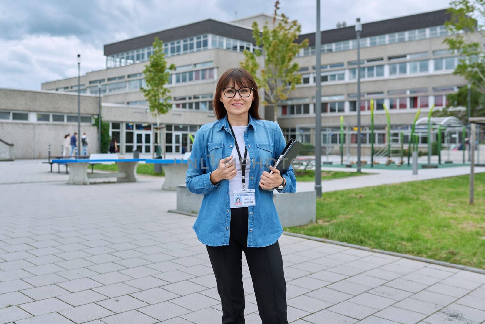Middle-aged confident woman school teacher, mentor, pedagogue, psychologist, counselor, social worker with digital tablet in hands posing near school building, outdoor. Education, teaching, school
