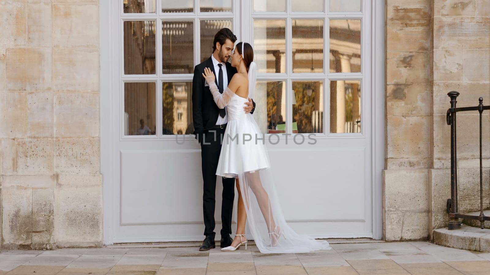 Happy newlyweds embracing each other by the white entrance door with windows. Action. Young and pretty man and woman in suit and white short dress