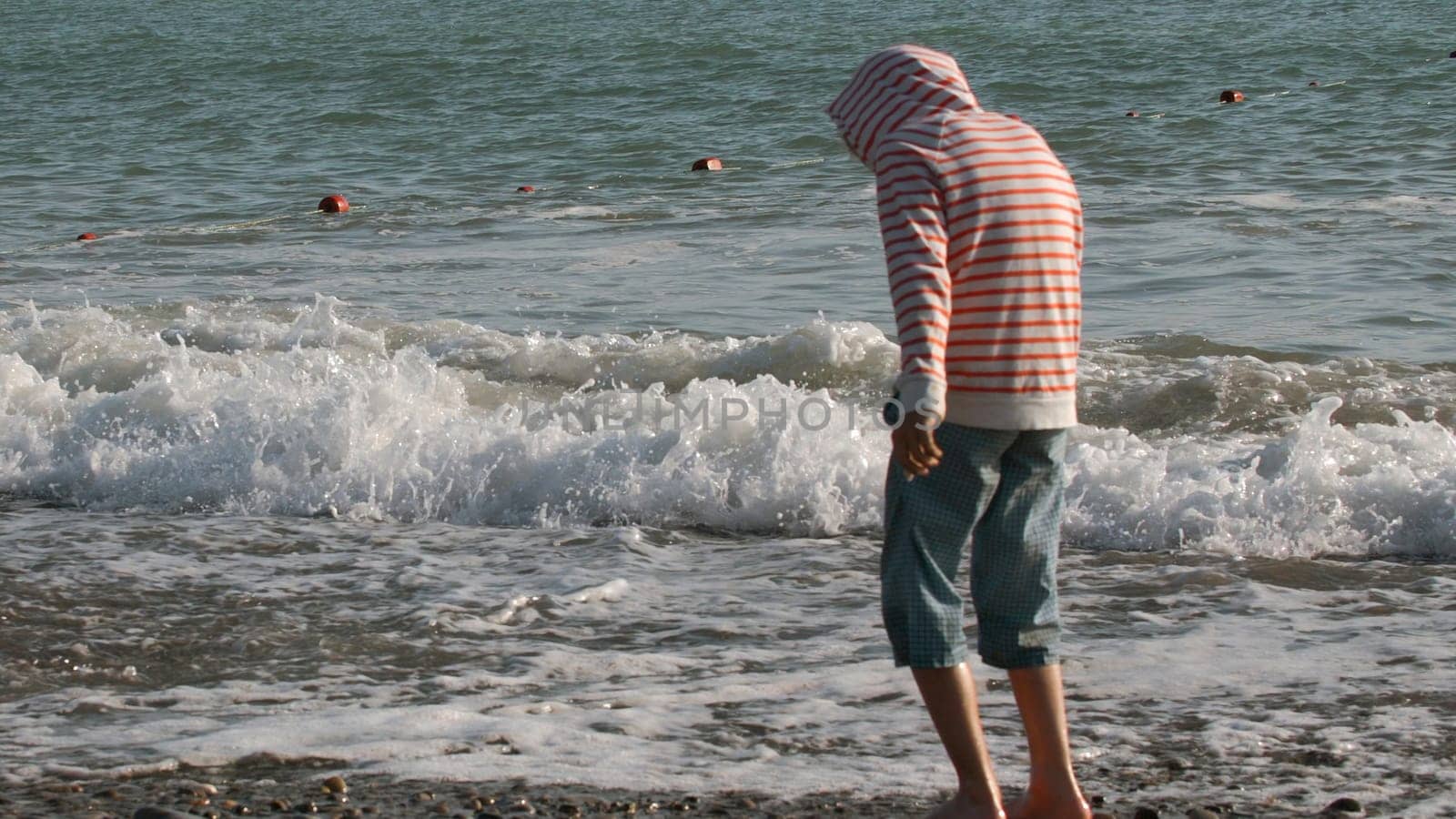 Young boy standing barefoot in the water on the pebble shore. Creative. Rear view of a boy exploring sea shore