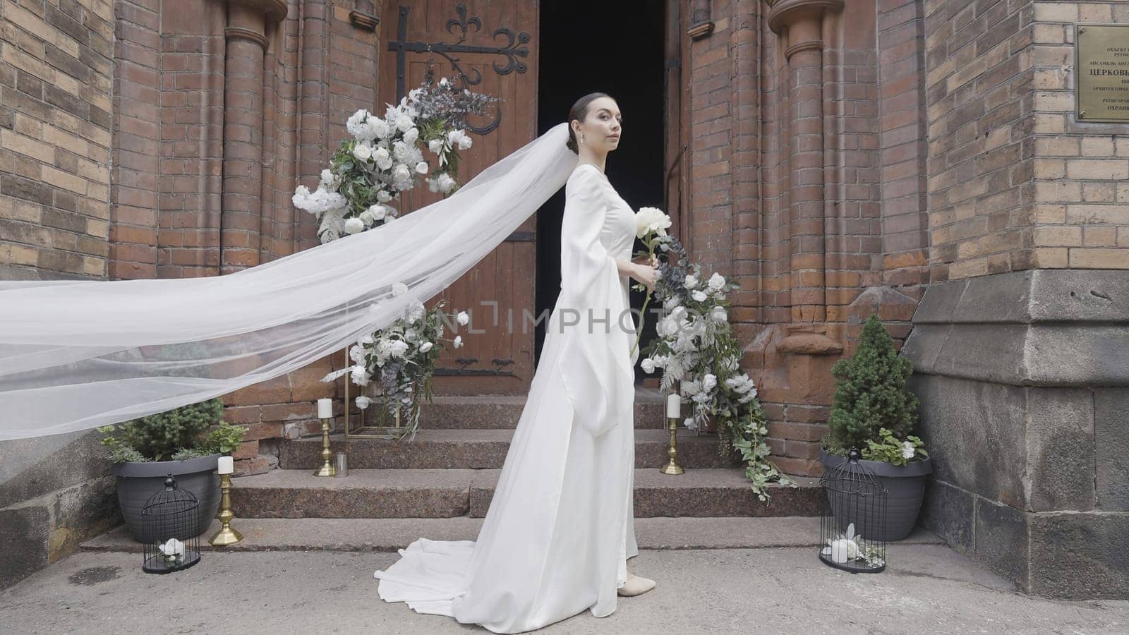 Elegant bride at entrance to church. Action. Luxurious bridal veil with bouquet at church. Beautiful bride's outfit with long veil.