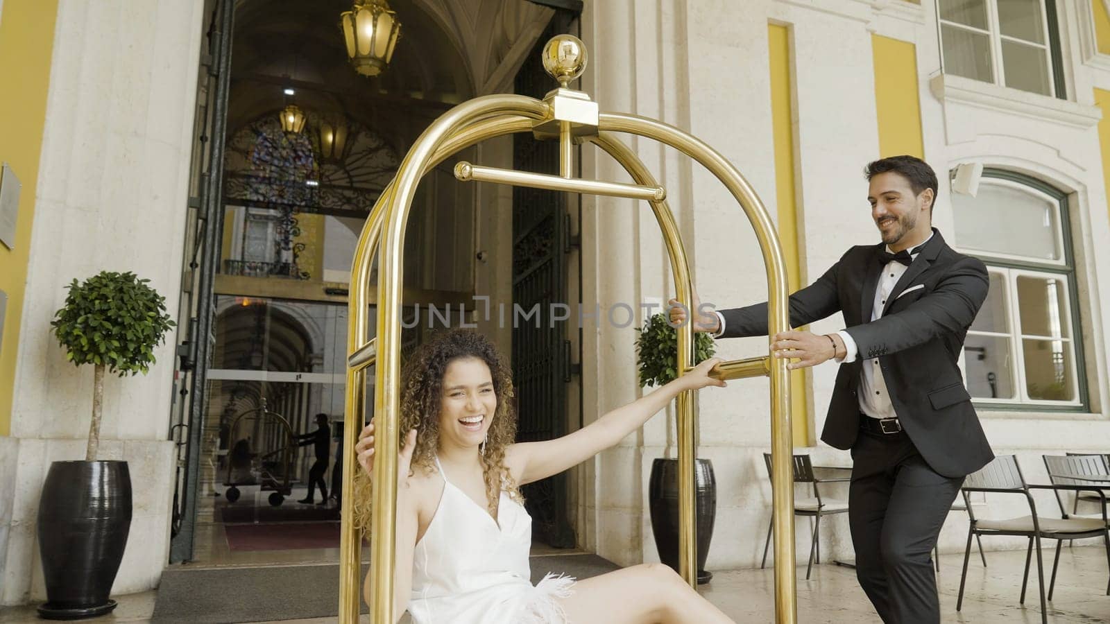 Groom riding bride in luggage trolley near luxury hotel. Action. Newlyweds having fun