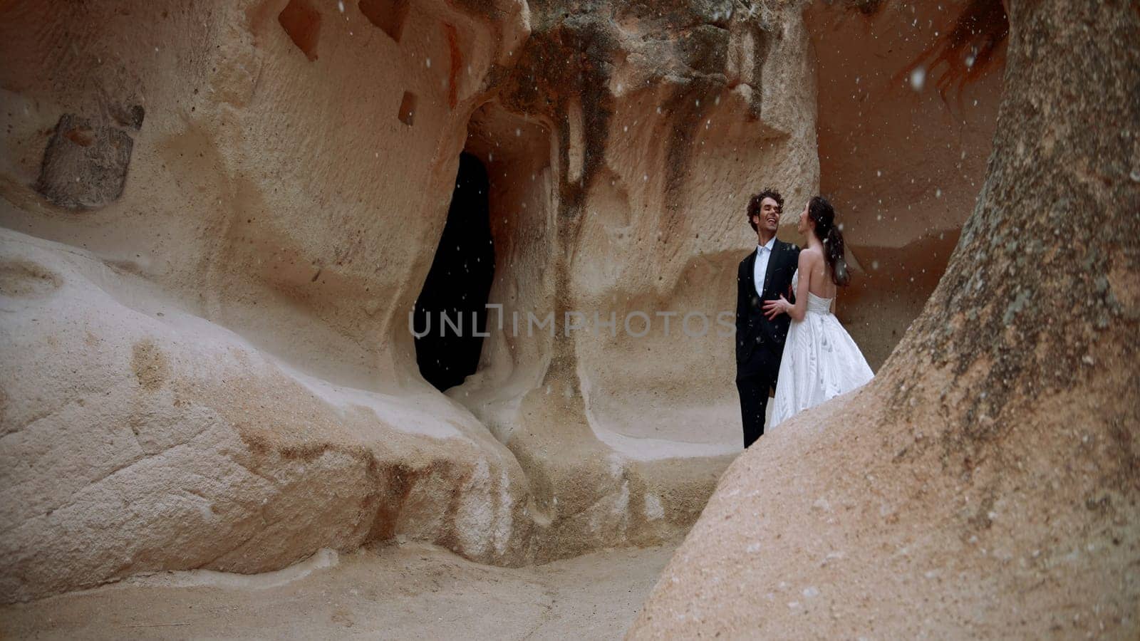 Stylish beautiful bride and groom under falling snow near rocks or caves. Action. Wedding video shooting in cappadocia. by Mediawhalestock