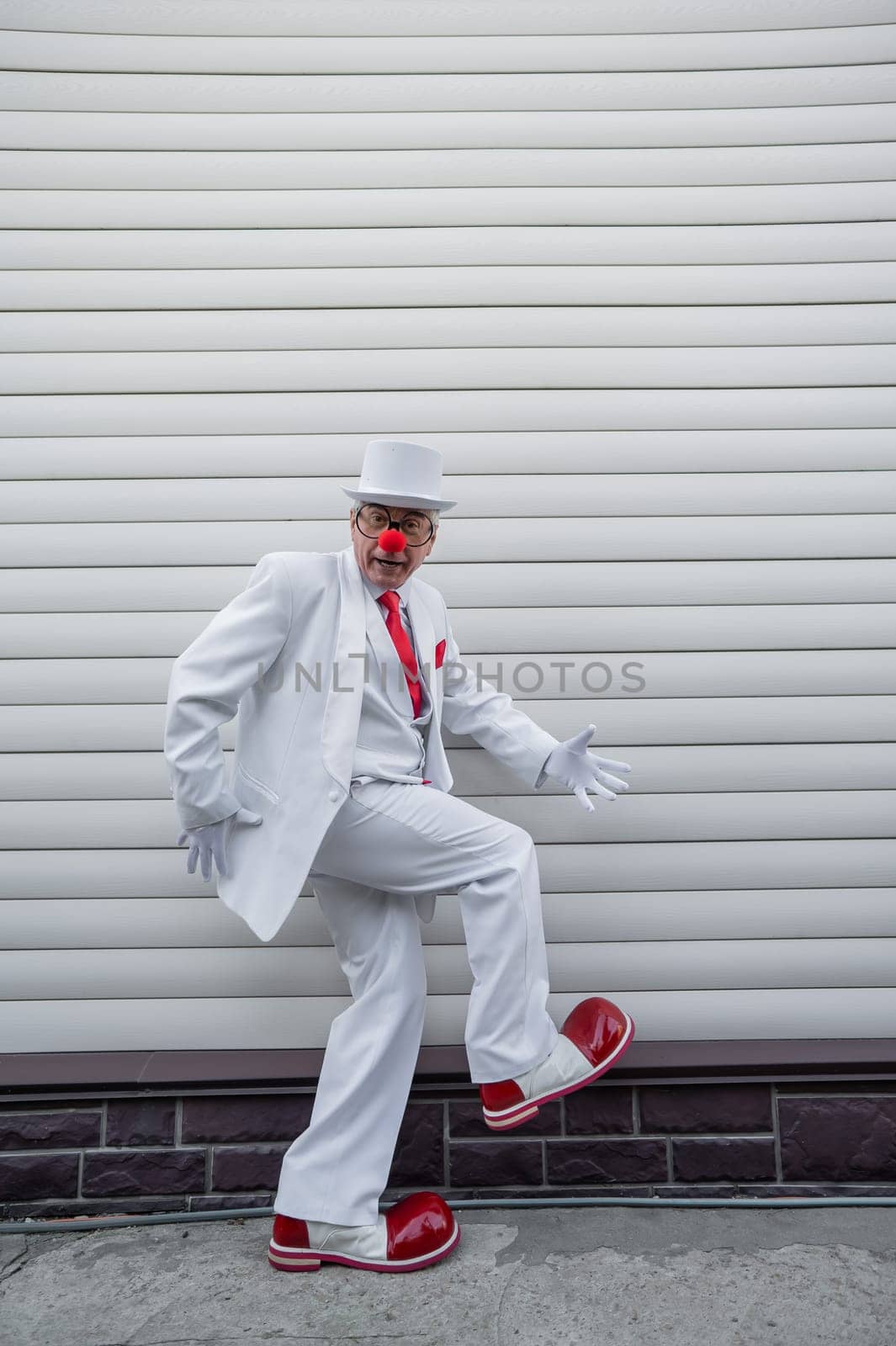 An elderly man in a white suit, huge boots and a clown nose walks funny. by mrwed54