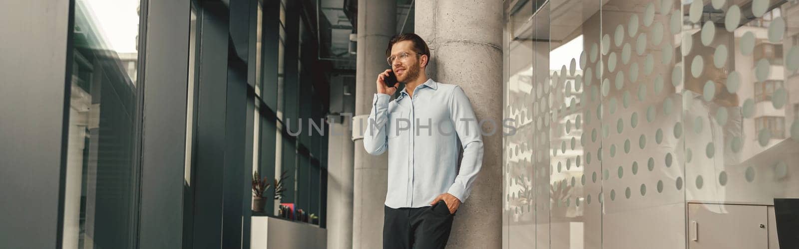 Smiling male sales manager talking phone with client while standing in coworking and looking at side by Yaroslav_astakhov