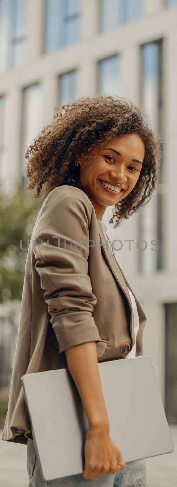 Smiling businesswoman standing with laptop on office building background during break time by Yaroslav_astakhov