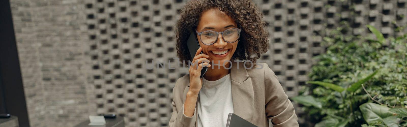 Smiling businesswoman is talking phone with client while standing on office background by Yaroslav_astakhov