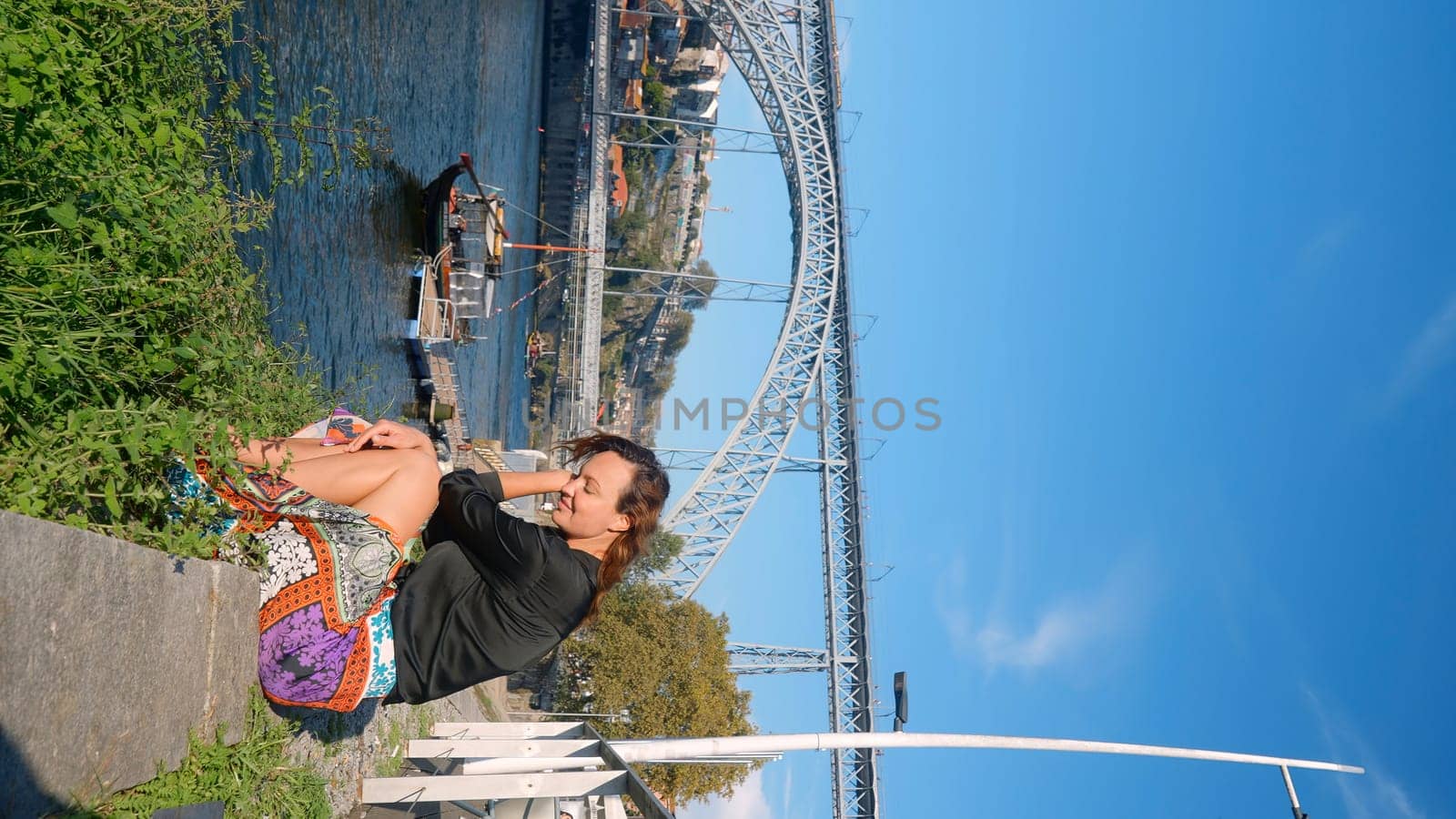 Vertical video with beautiful woman on embankment. Action. Attractive woman is sitting on edge of embankment on sunny summer day. Stylish woman is resting on city embankment with view of bridge.