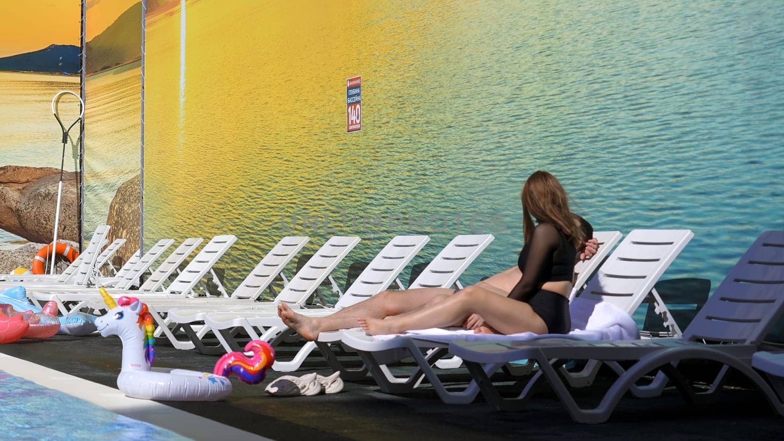 Young man joining to his beautiful brunette girlfriend on sunbed. Clip. Concept of relax, summer vacation