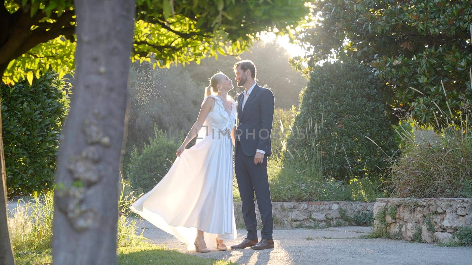 Happy bride and groom walking and holding hands in the park. Action. Wedding couple during video shooting among green summer vegetation