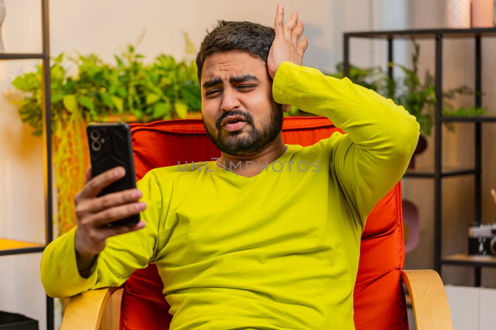 Sad displeased Arabian man use smartphone typing browsing, loses becoming surprised sudden lottery game results, bad news, fortune loss fail, deadline, virus. Young Indian guy at home room on chair