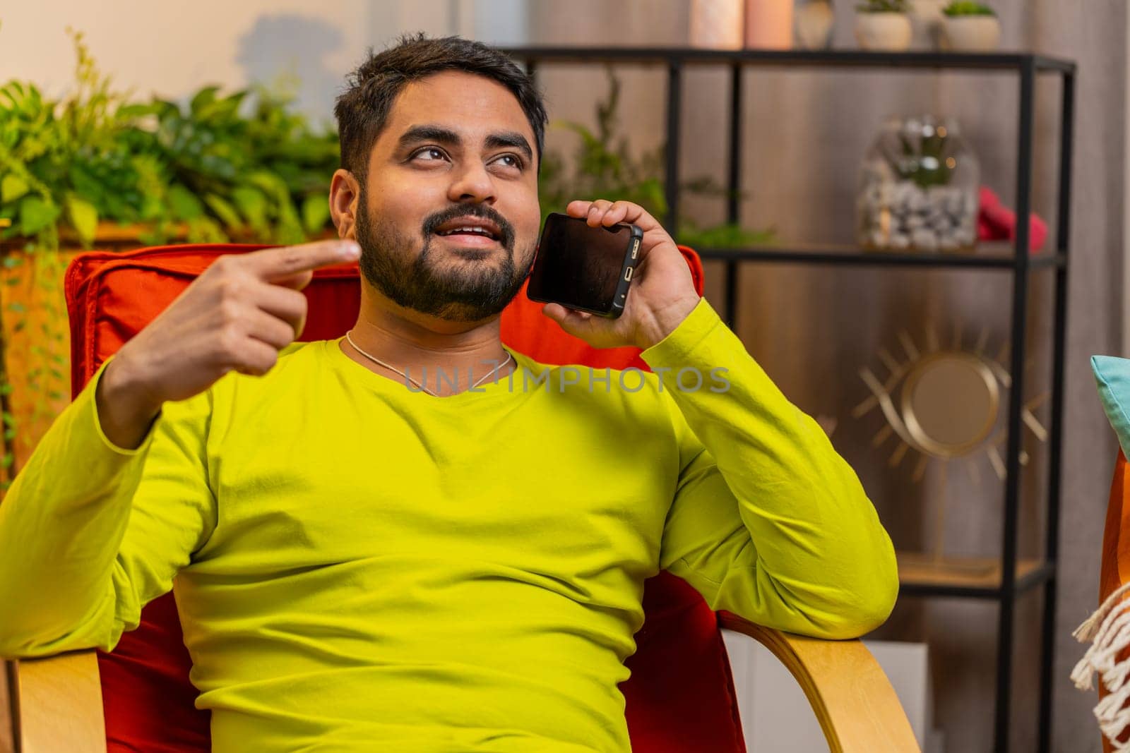 Phone call, good news, gossip. Happy surprised Indian man having pleasant conversation on smartphone, enjoying talking with friend at modern home room. Arabian guy enjoying mobile phone speaking