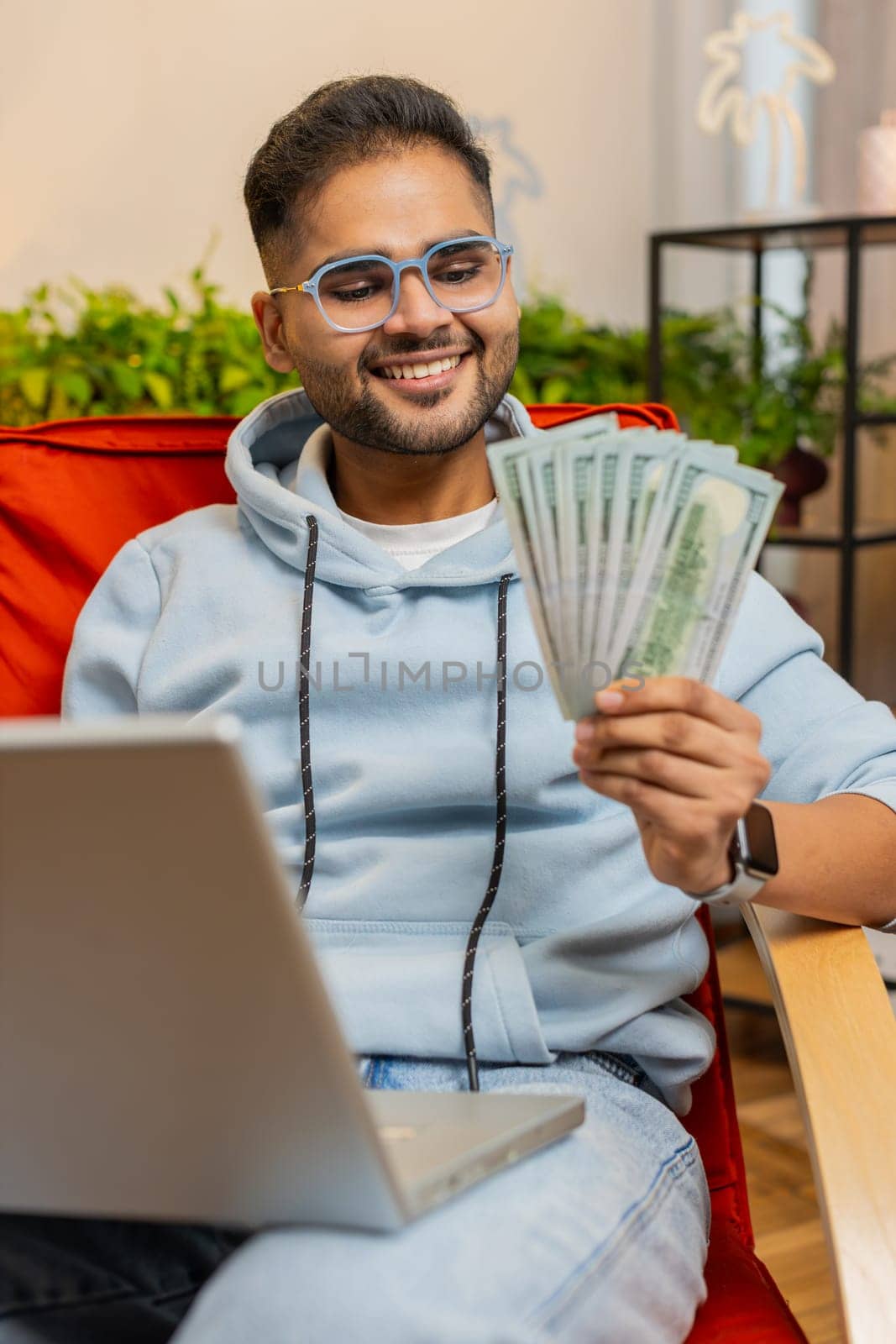 Planning budget. Rich happy Indian man counting money cash use laptop computer calculate domestic bills at home. Arabian guy satisfied of income earnings, saves money for planned vacation, gifts