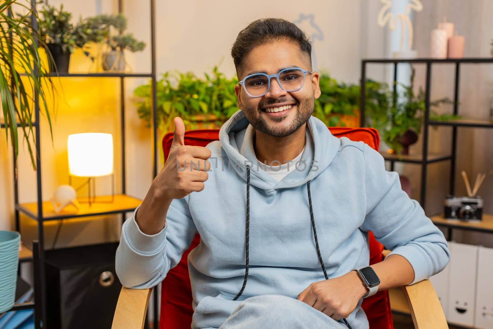 Like. Happy excited Indian man looking approvingly at camera showing thumbs up like sign positive something good great news positive feedback. Young Arabian guy sitting on chair at home living room