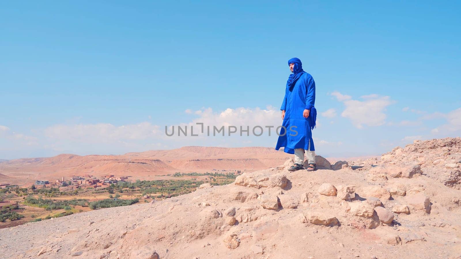 Dusty desert road with low growing bushes and plants on a blue background. Action. A man in blue male arabic clothes standing on a hill