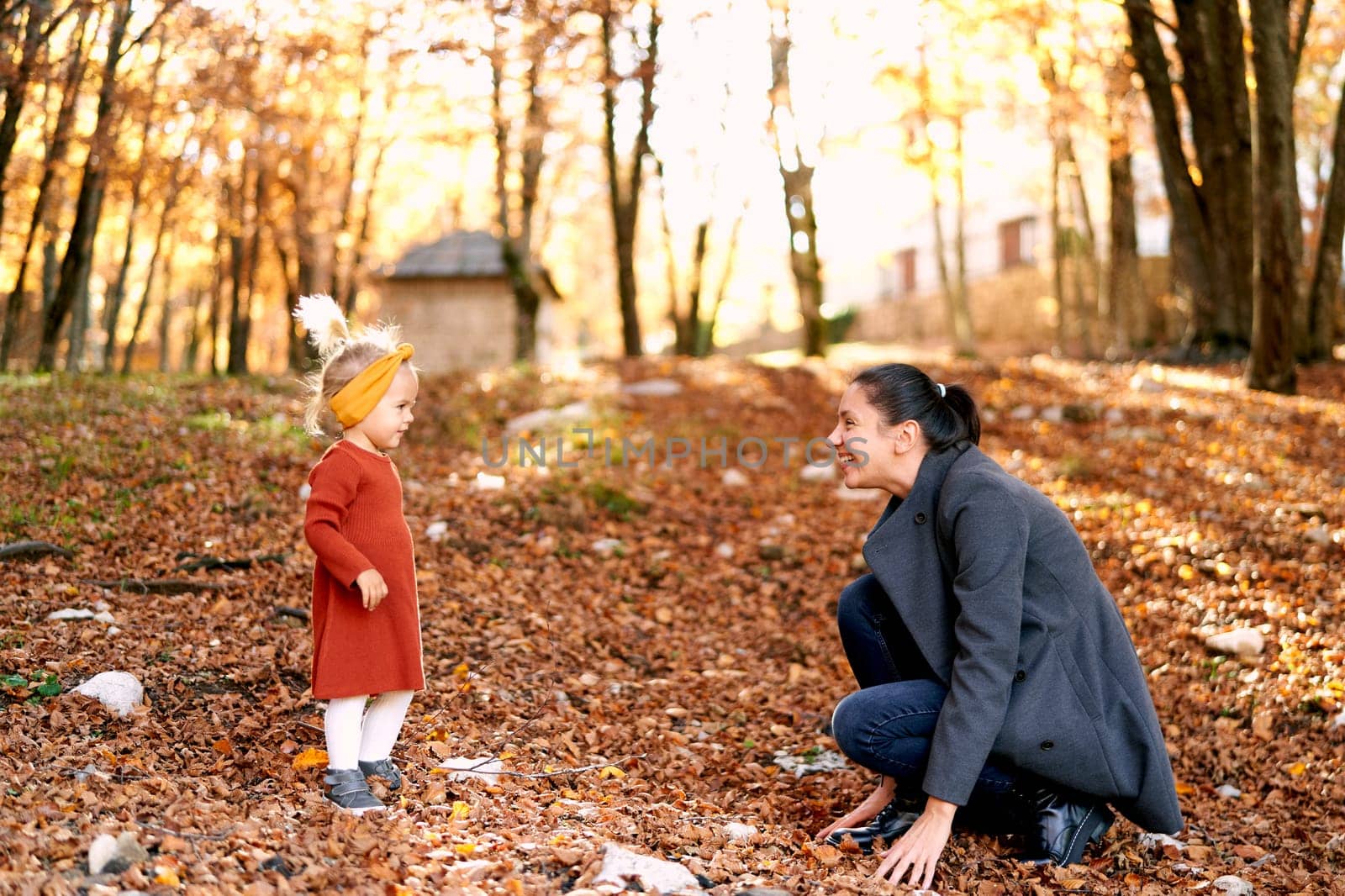 Little girl stands in front of her mother squatting in the autumn forest touching the fallen leaves by Nadtochiy