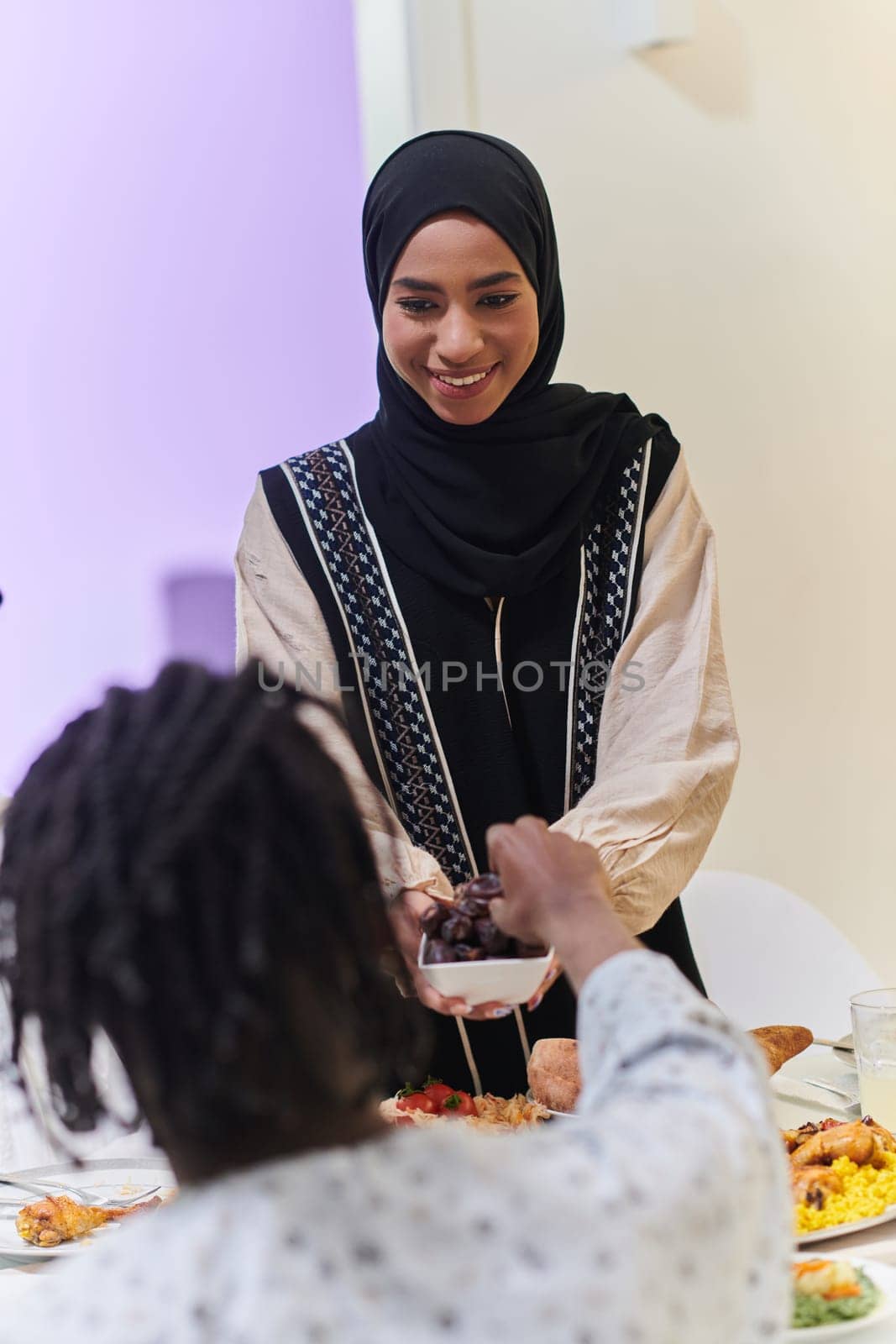 In a heartwarming scene during the sacred month of Ramadan, a traditional Muslim woman offers dates to her family gathered around the table, exemplifying the spirit of unity, generosity, and cultural richness during this festive and spiritual occasion by dotshock