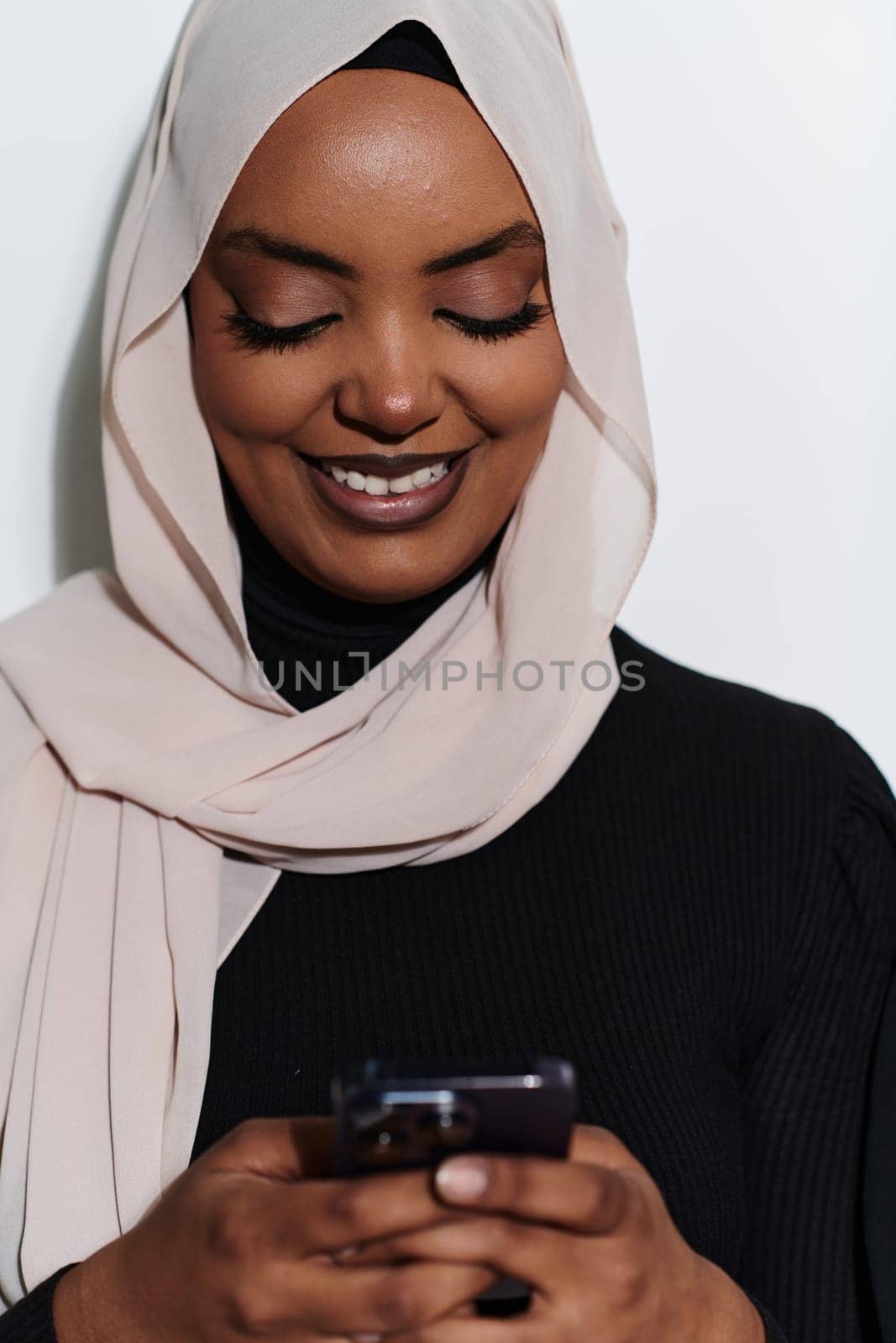 An elegant Arab woman, adorned in a hijab, engages with modernity as she uses a smartphone, the juxtaposition of traditional attire against contemporary technology captured in the isolated setting against a pristine white background.